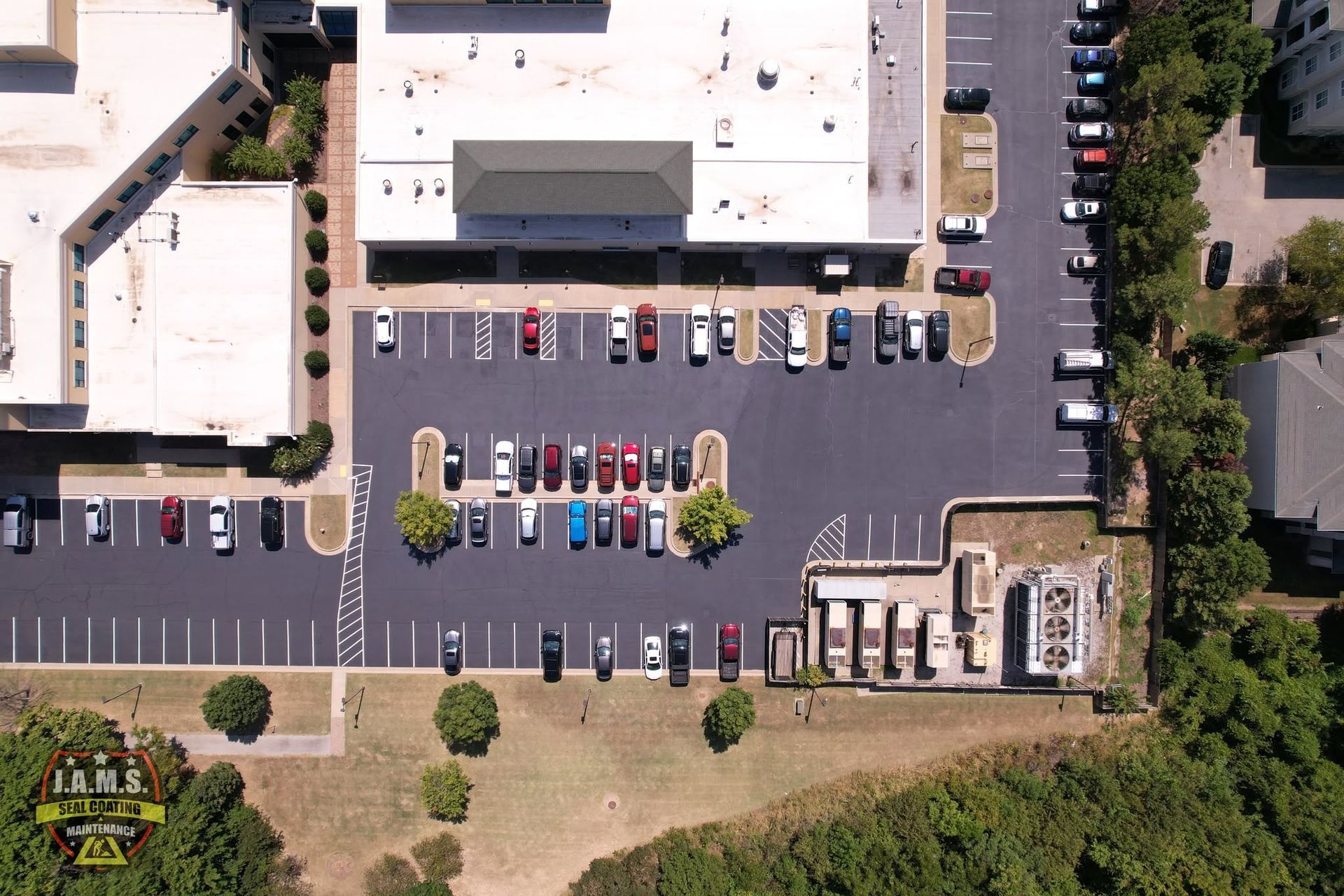 Aerial view of a commercial parking lot with parked cars beside a large building and trees.