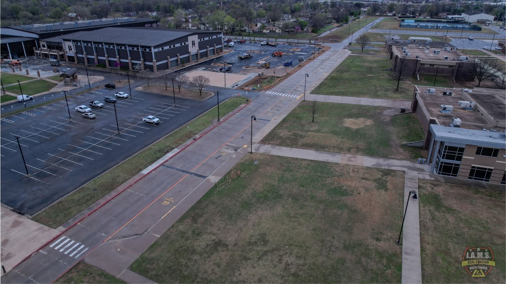 Aerial view of a road beside a parking lot, grassy field, and low buildings on an overcast day