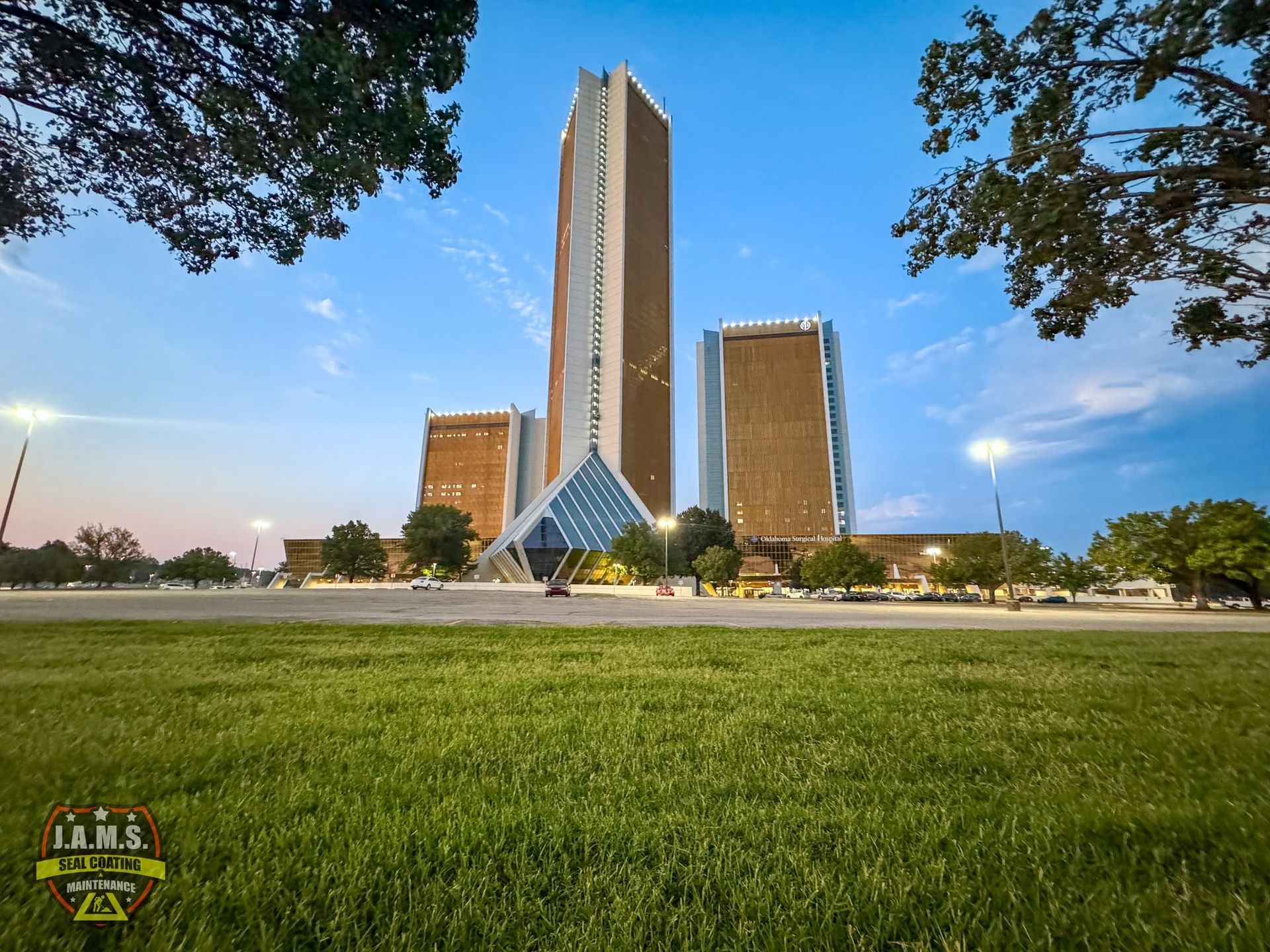 Tall city building with a blue glass pyramid entrance, framed by trees and a grassy lawn at dusk