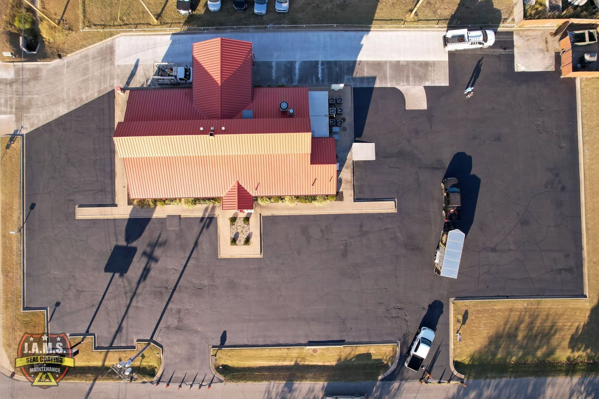 Aerial view of a small building with a red roof and parking lot, with a few vehicles and shadows nearby.
