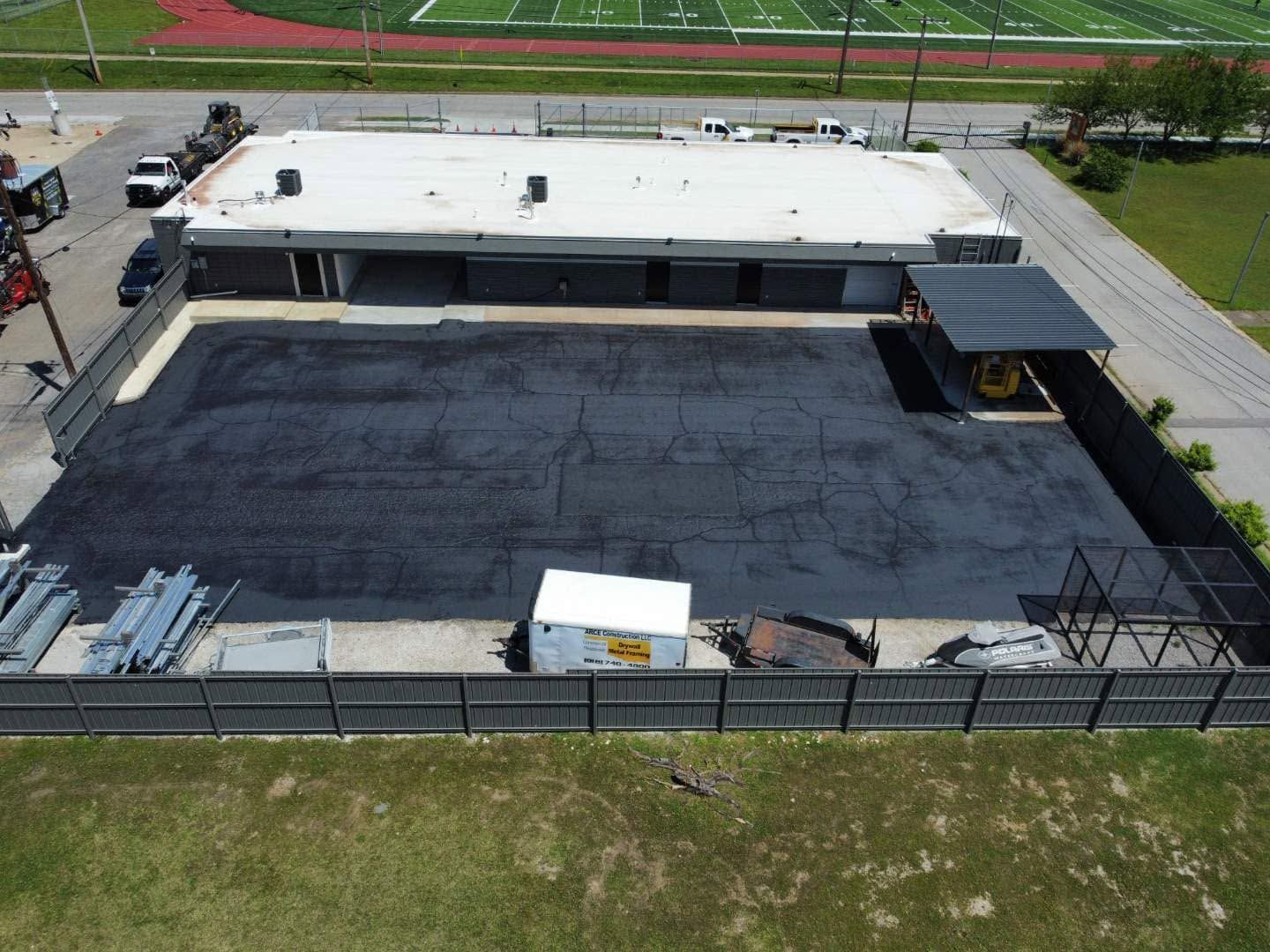 Aerial view of an empty paved lot beside a long white building and fenced grassy field.