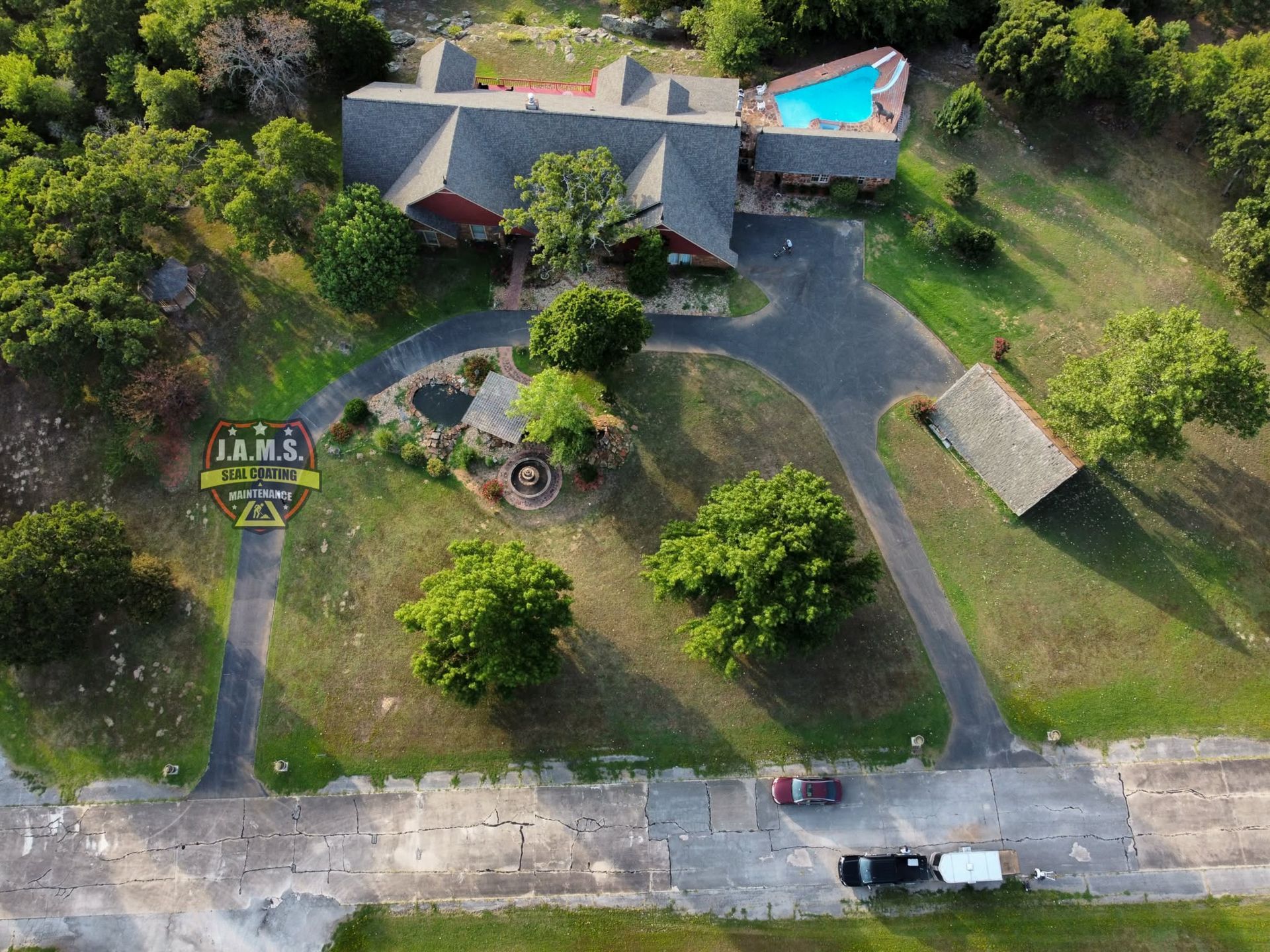 Aerial view of a rural house with a driveway, green yard, trees, and a parked vehicle by the road