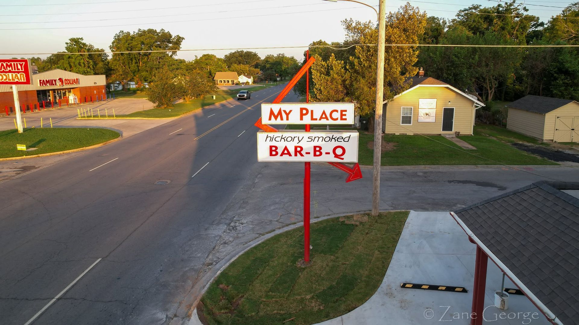 Roadside sign for My Place Bar-B-Q beside a quiet street and small buildings at dusk