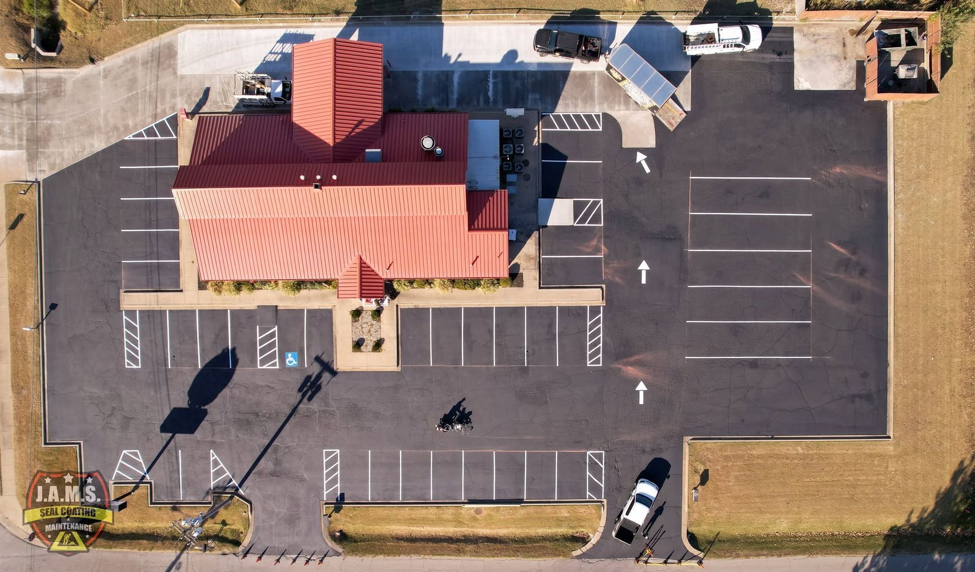 Aerial view of a small building with a red roof beside an empty parking lot and marked spaces