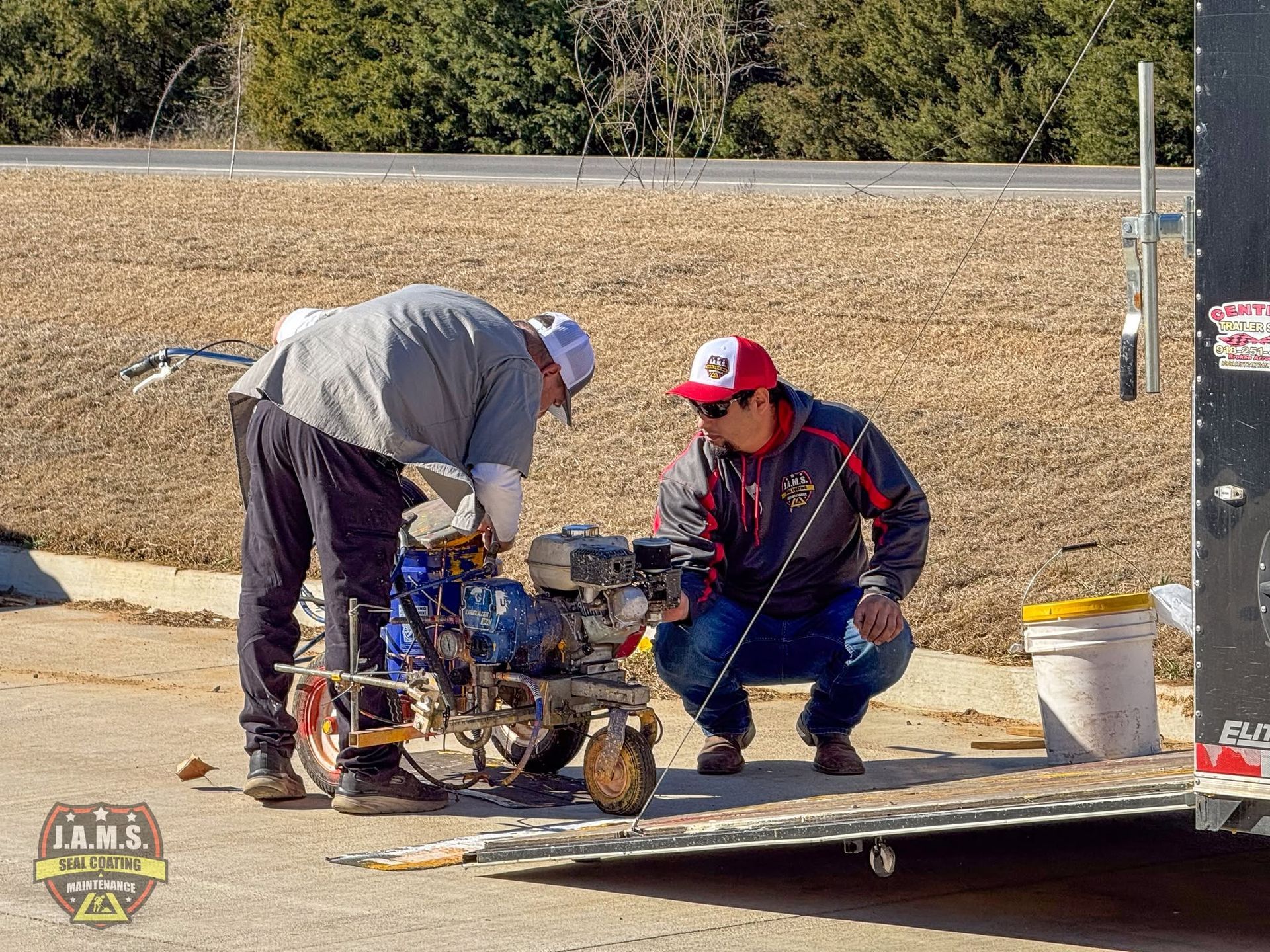 Two workers crouch by a small engine on a flatbed trailer in an outdoor gravel lot.