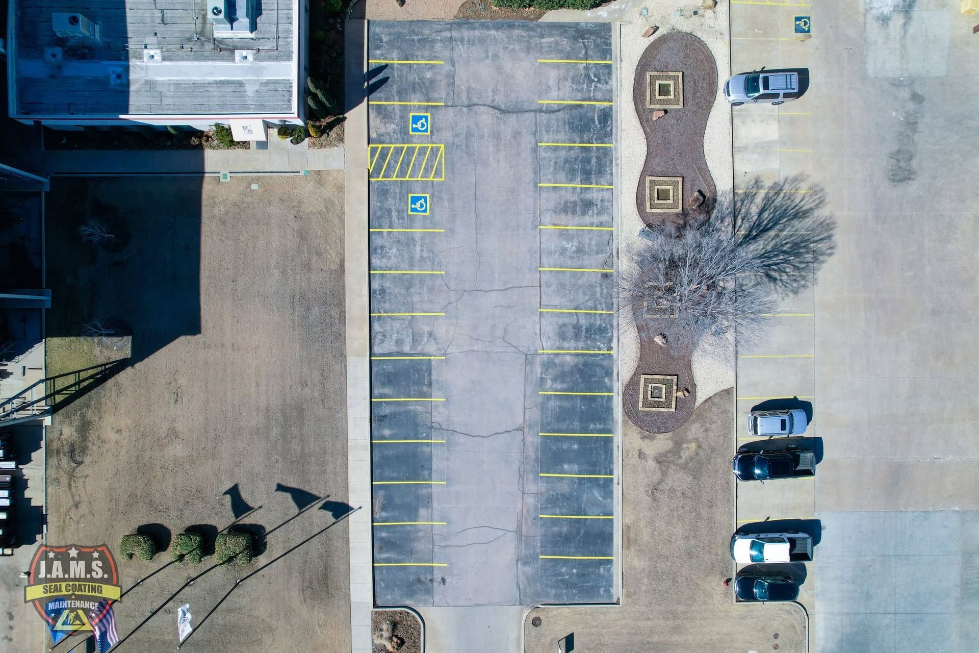 Aerial view of an empty parking lot with marked spaces, a few cars, and a tree on the right.