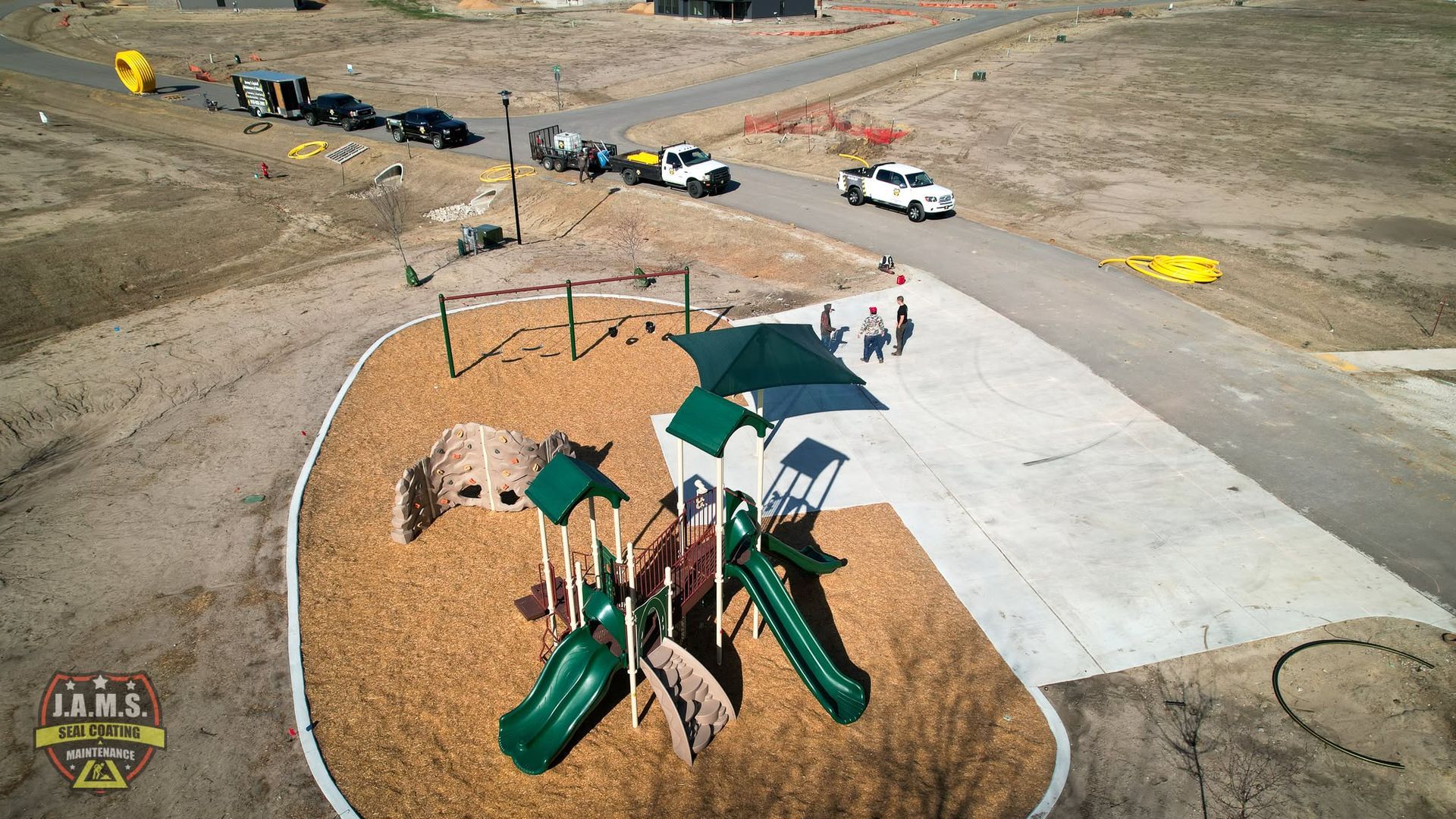 Aerial view of a playground with green slides and tan surfacing beside a parking lot and dirt road.