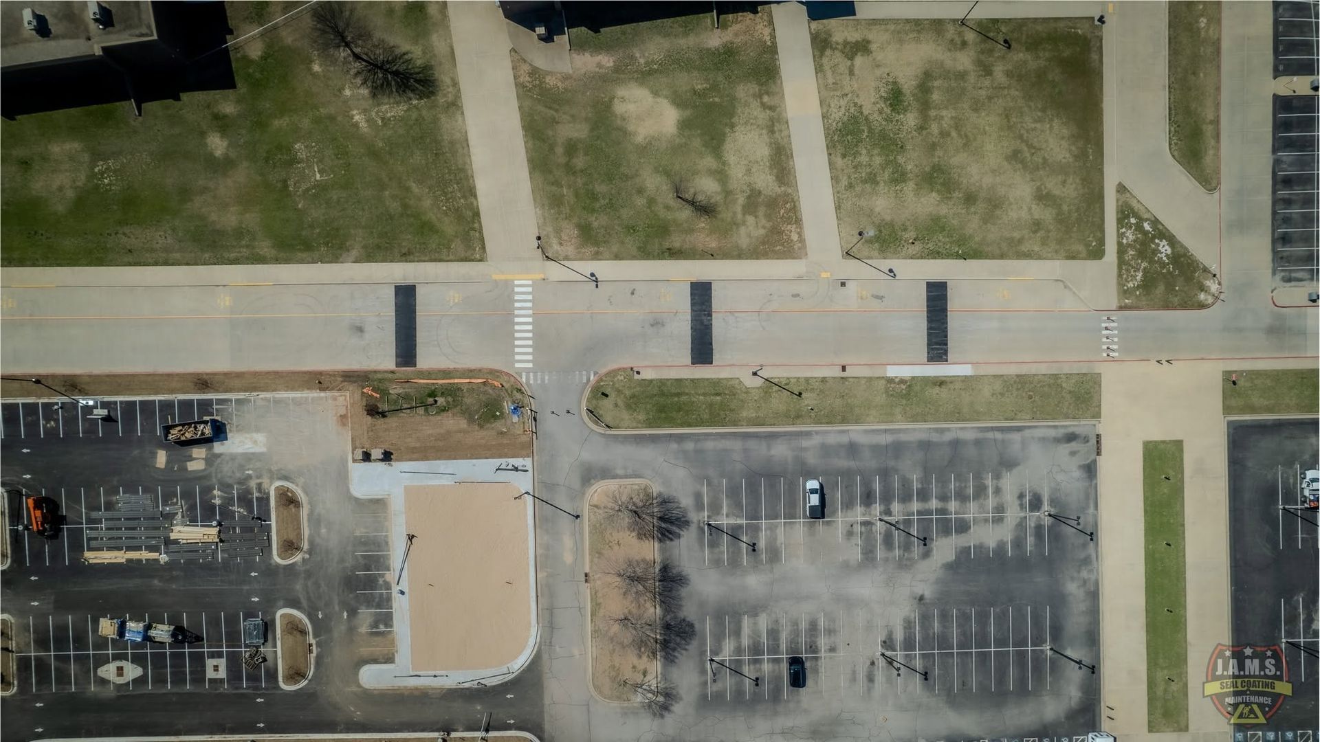 Aerial view of a parking lot, road, and grassy plots with a few parked cars and buildings nearby
