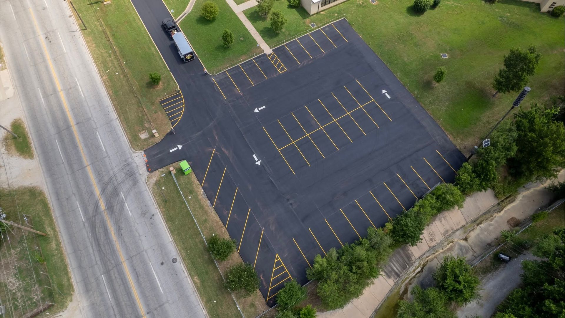 Aerial view of an empty blacktop parking lot with yellow lines beside a road and grassy trees.