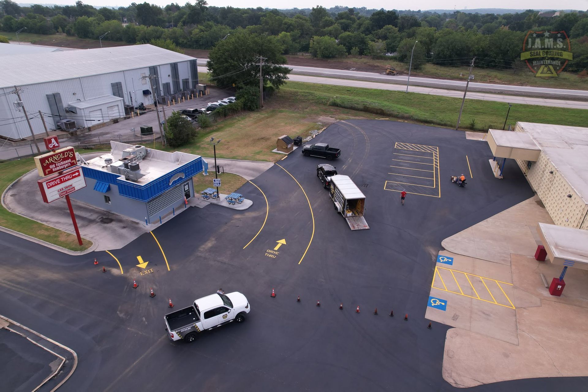 Aerial view of a parking lot with a blue-roofed kiosk, trucks, cones, and a large warehouse building.