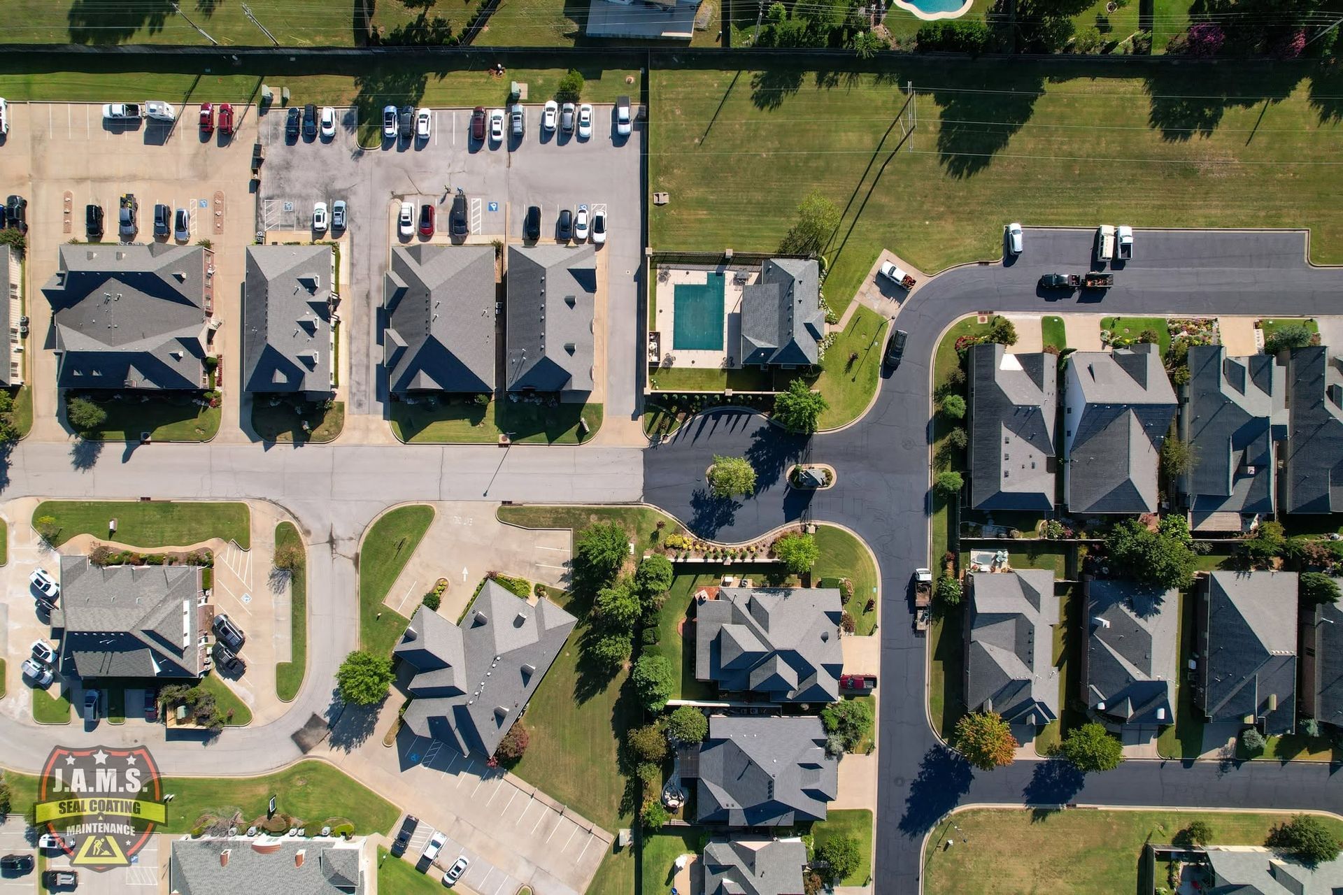 Aerial view of a suburban apartment complex with parked cars, roads, lawns, and a small pond.