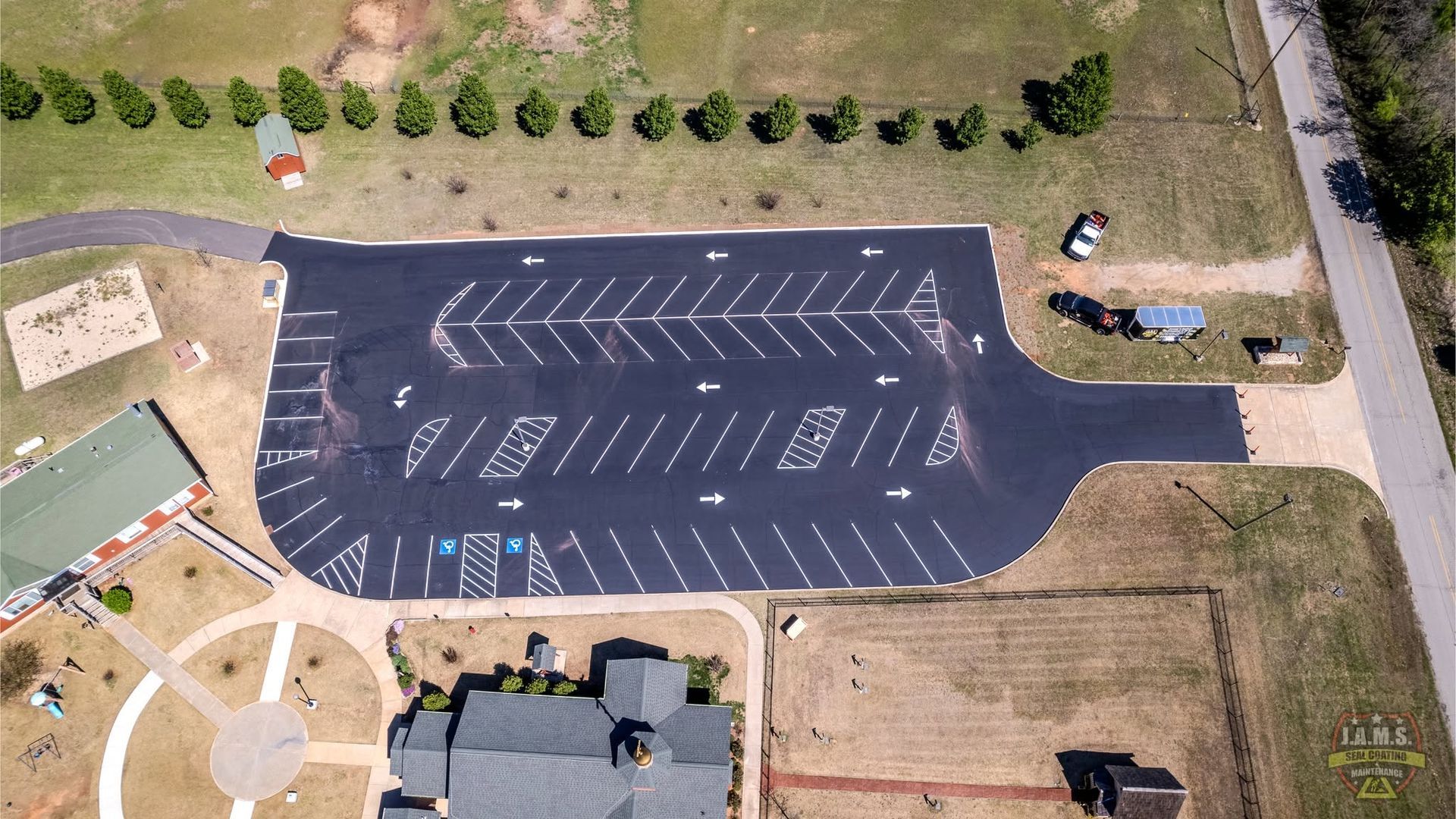 Aerial view of a mostly empty paved parking lot with marked spaces, trees, and nearby buildings and dirt lots
