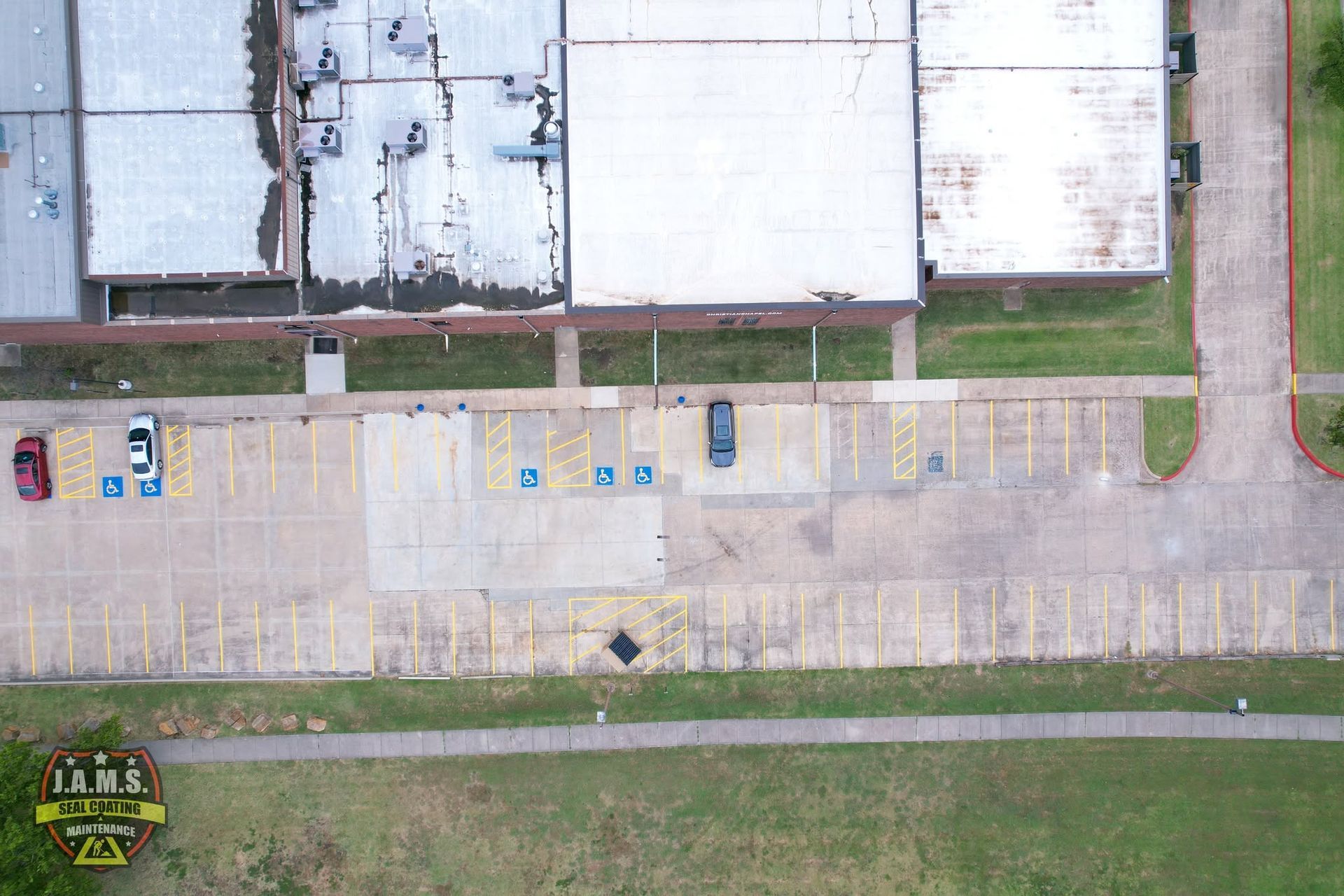 Aerial view of a mostly empty parking lot beside large buildings, with a few cars and grass borders.