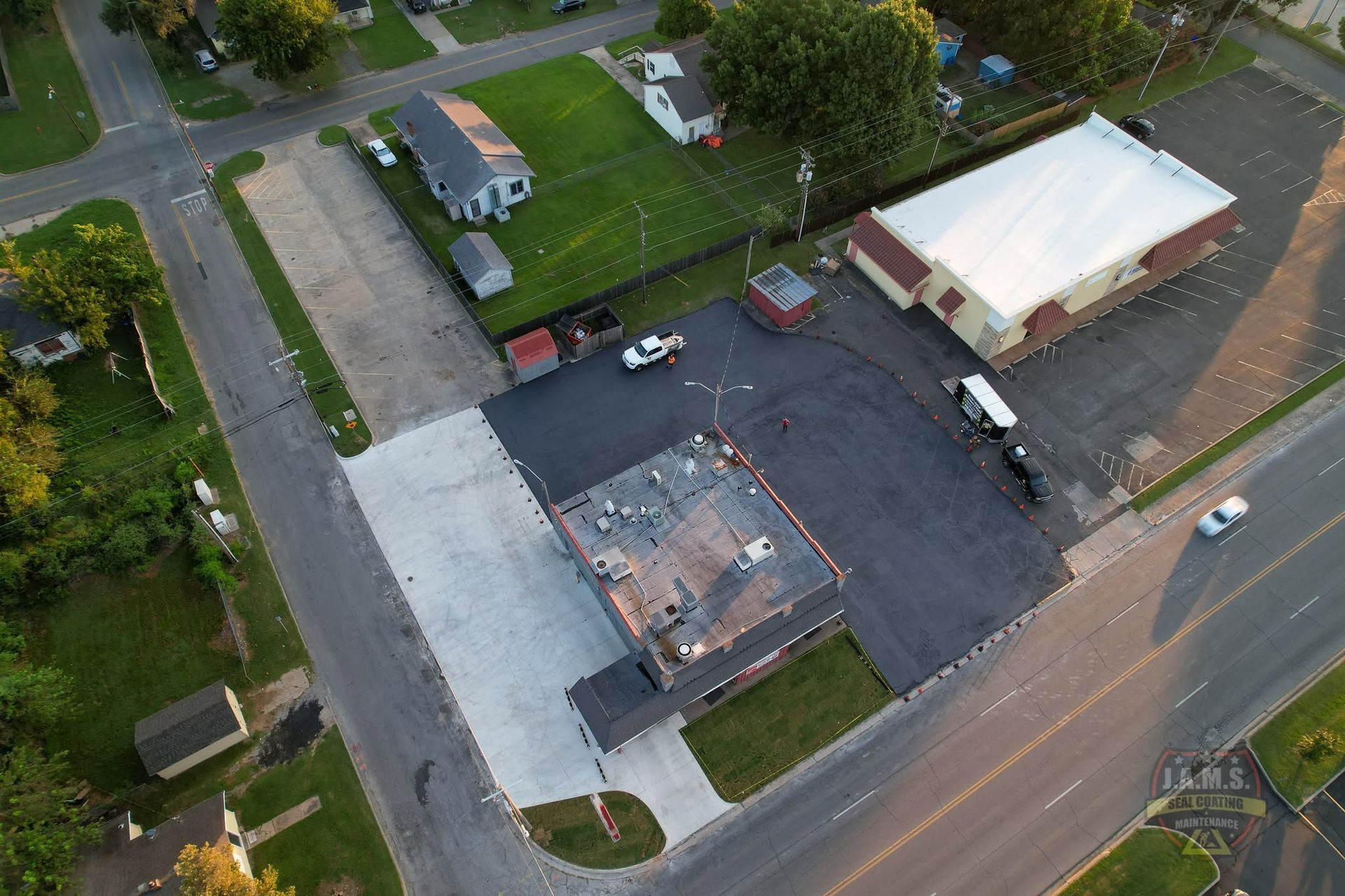 Aerial view of a small parking lot beside two buildings and a road, with cars and surrounding trees.
