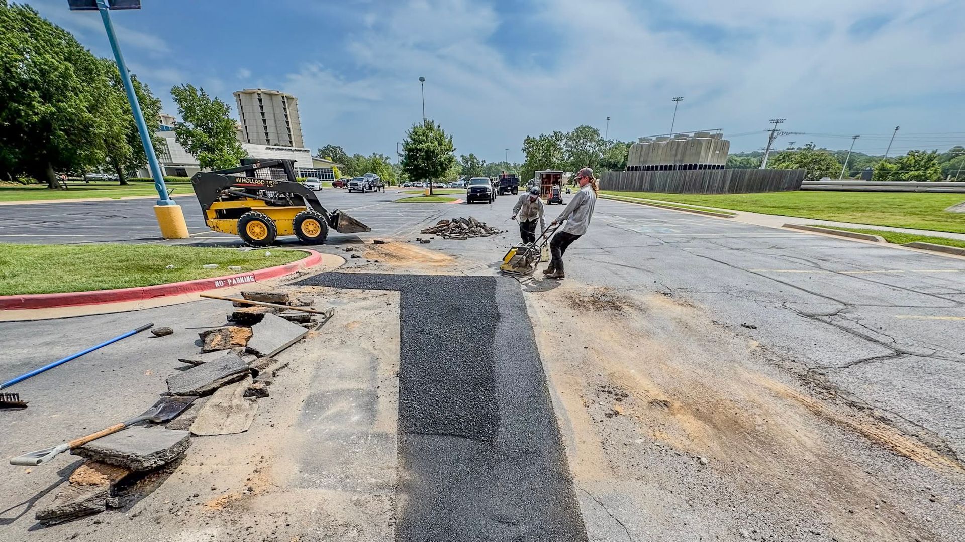 Roadwork scene with a partially torn-up road, workers, and a skid steer on a sunny street
