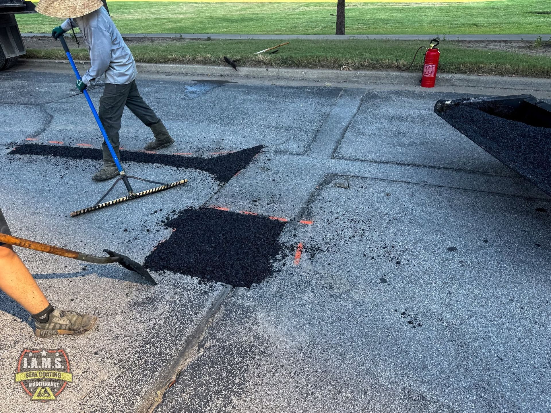 Workers patching a road with black asphalt near a grassy roadside, with cones and tools visible