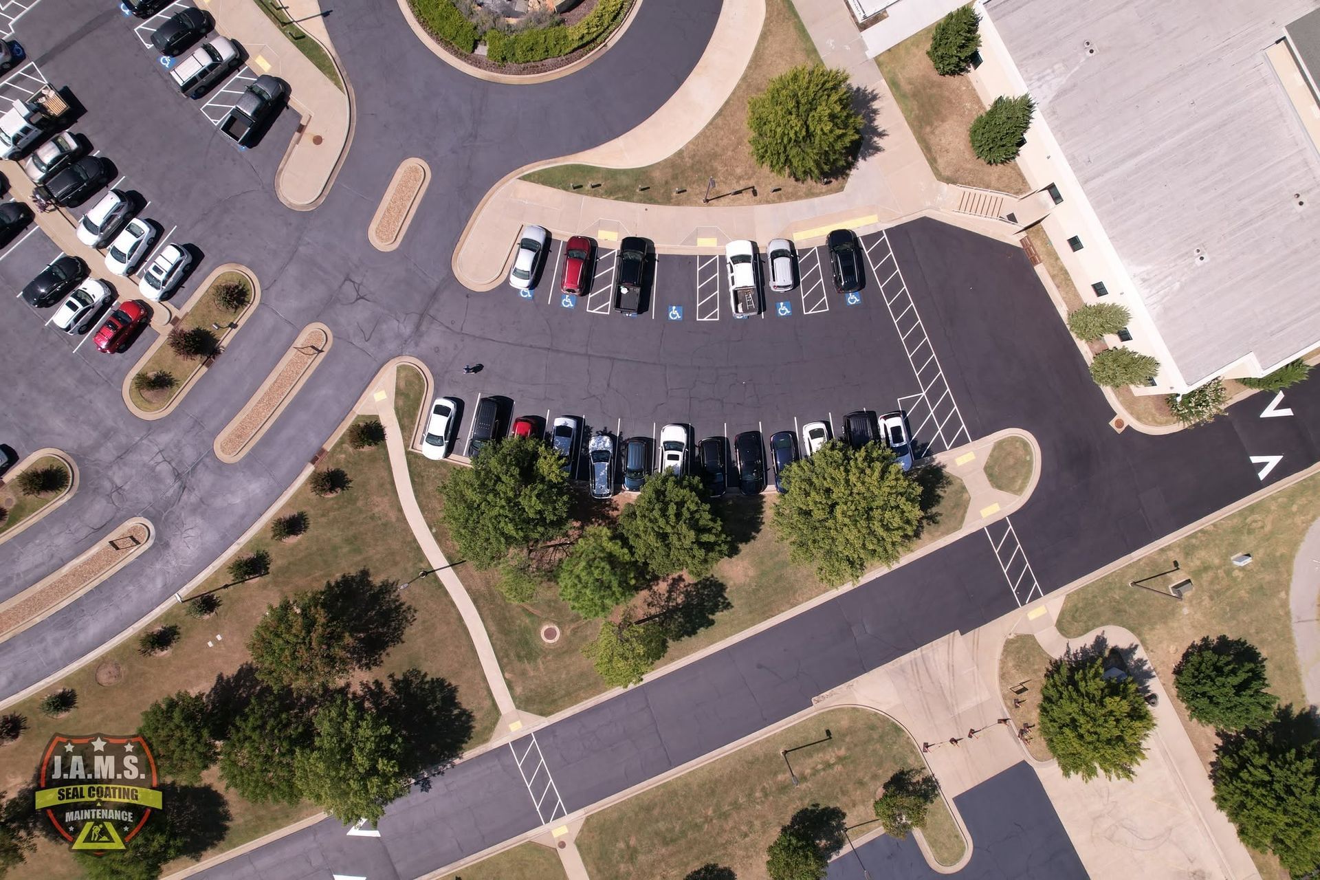 Aerial view of a parking lot with parked cars, trees, sidewalks, and surrounding buildings.