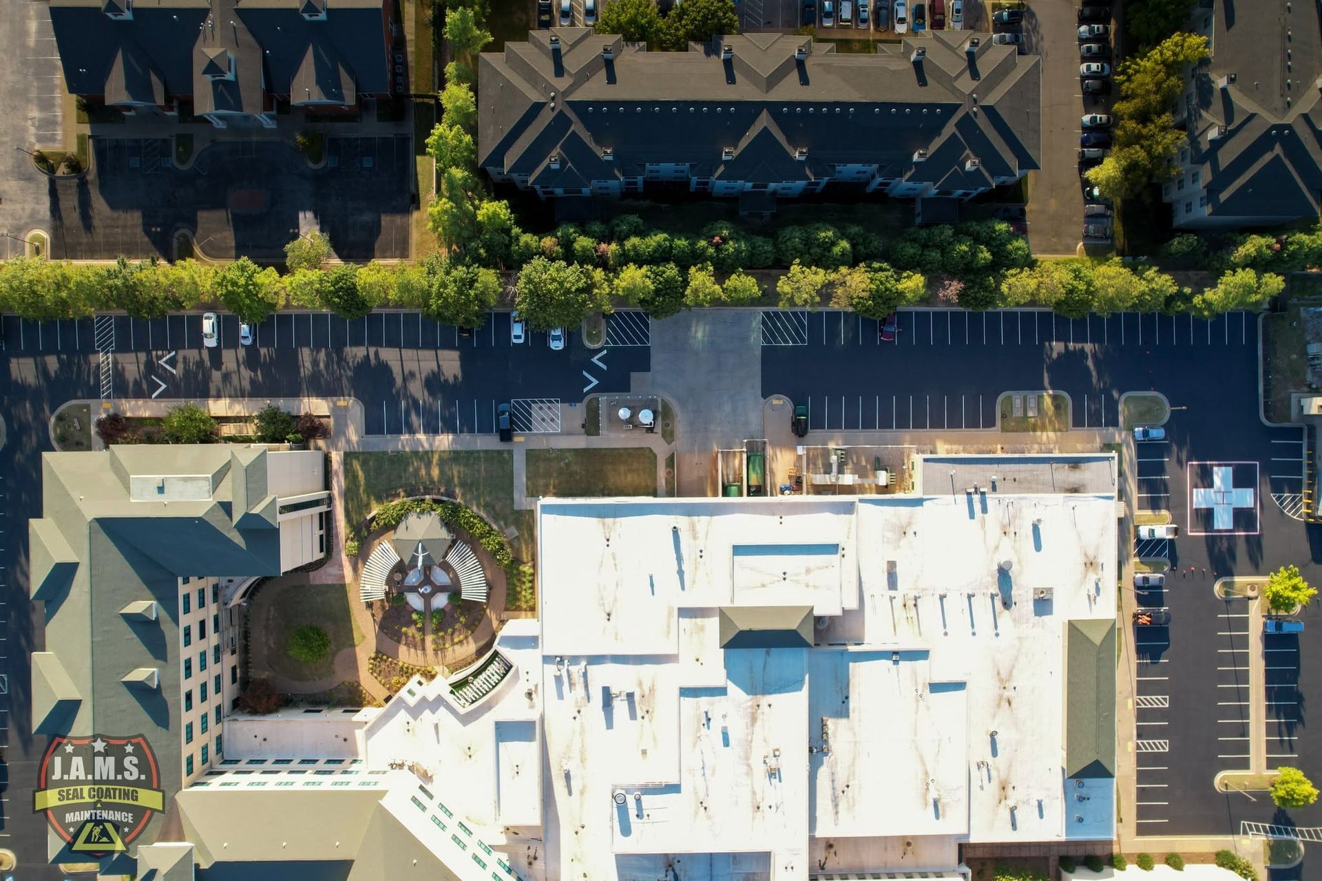 Aerial view of a parking lot beside white-roofed buildings and a tree-lined street.