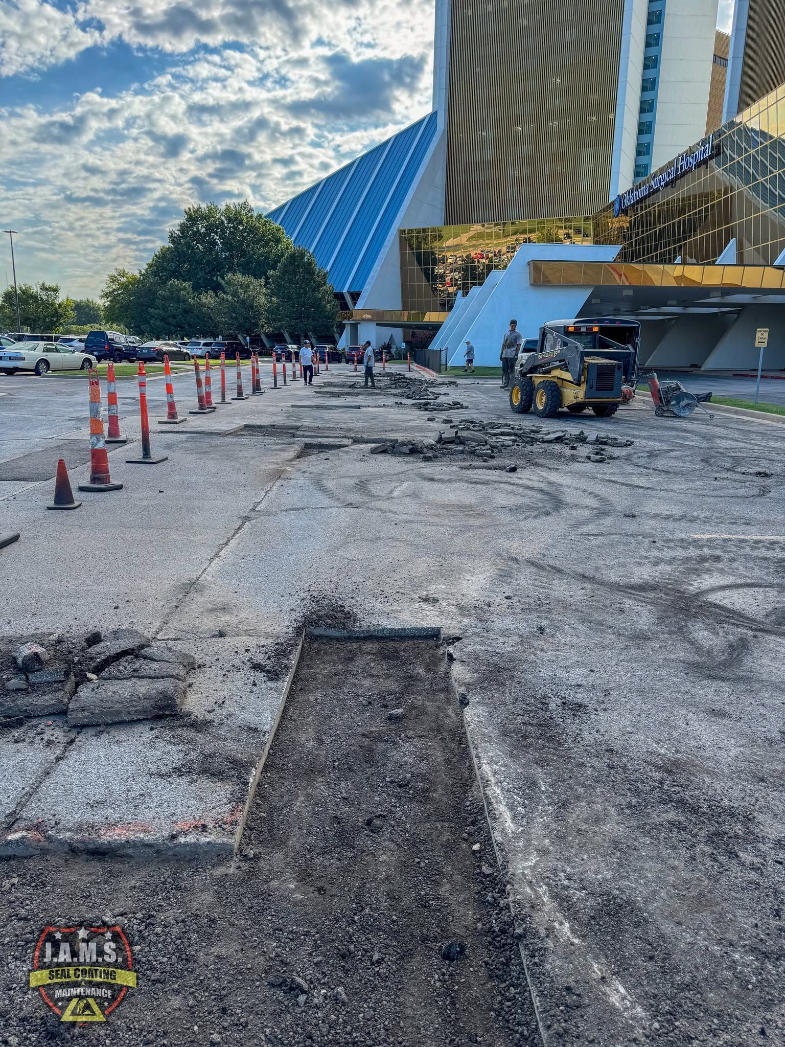 Construction work on cracked pavement outside a modern building, with cones and a backhoe near a fountain