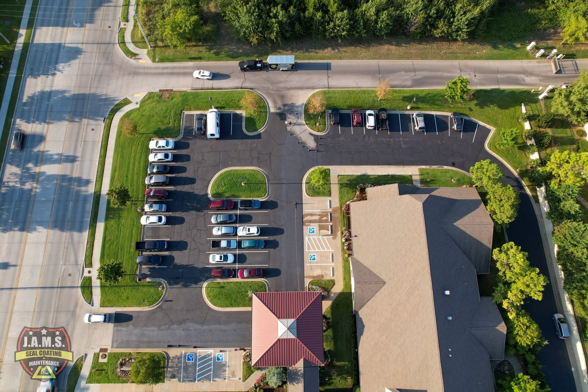 Aerial view of a parking lot beside a building and road, with several parked cars and trees.
