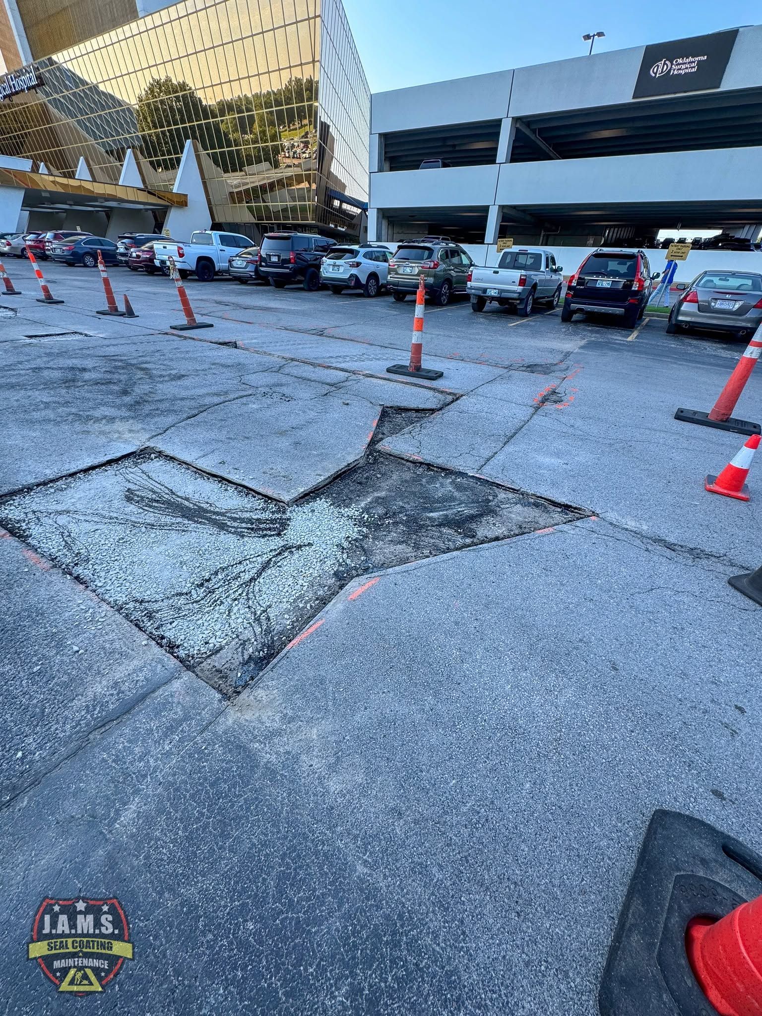Broken asphalt in a parking lot, bordered by orange cones near parked cars and a building.