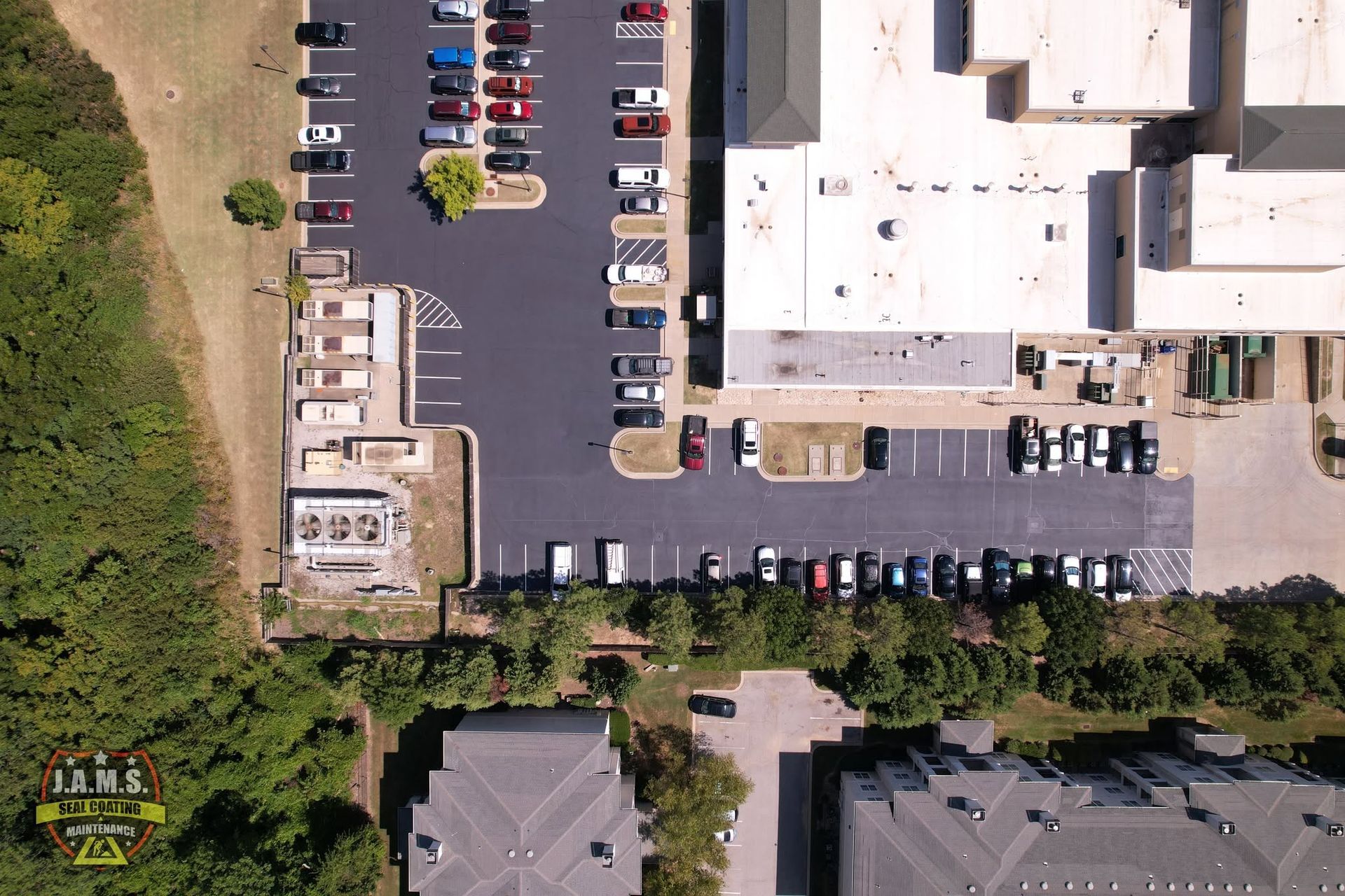 Aerial view of a parking lot beside a large white building and trees, with many parked cars.