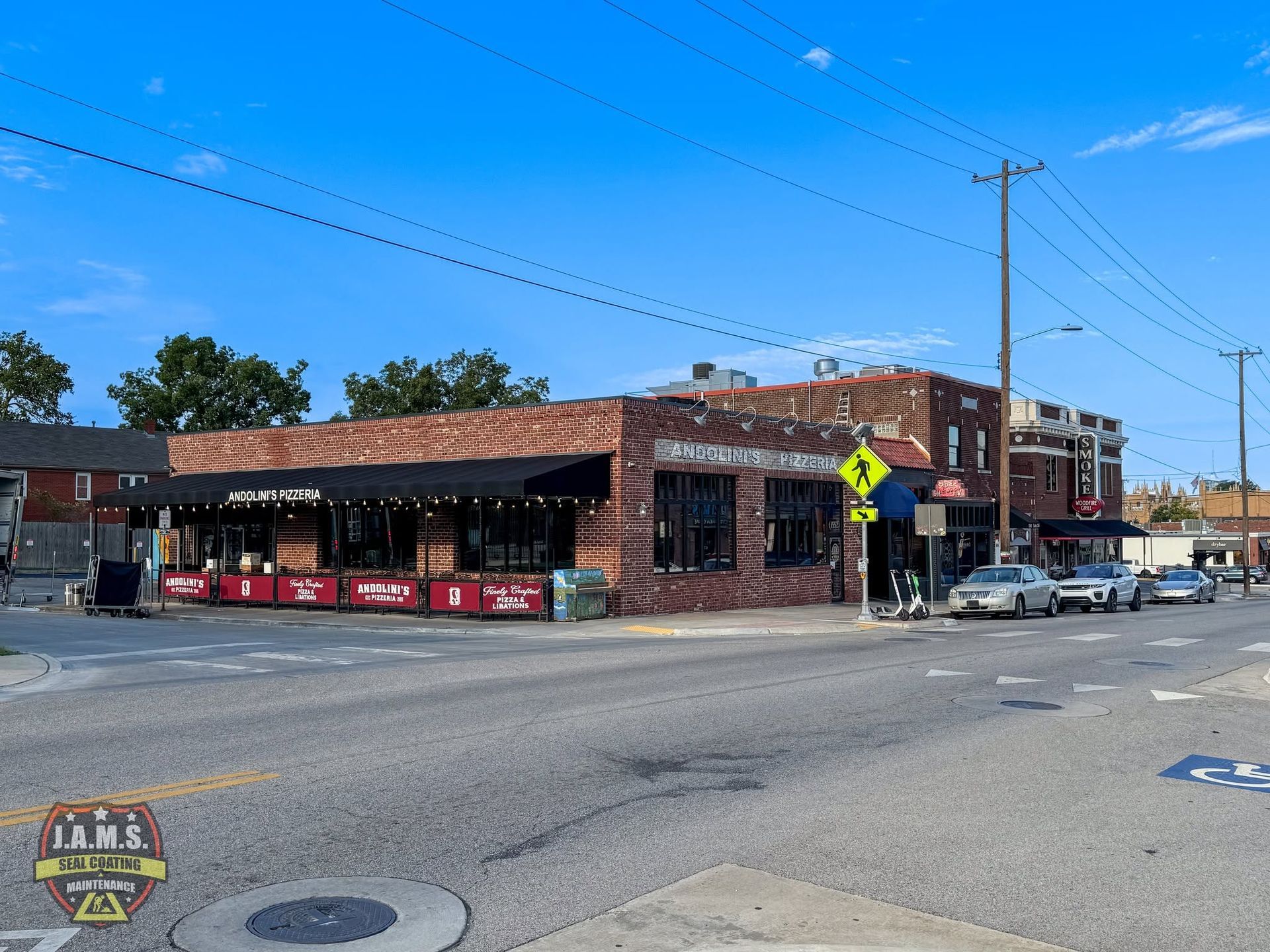 Brick corner storefront with a red awning, parked cars, and a pedestrian crossing sign on a sunny street.