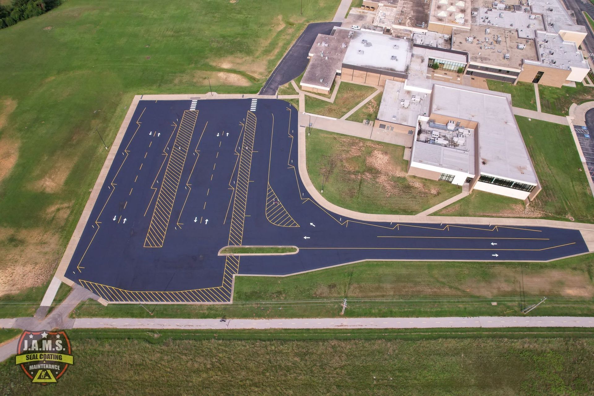 Aerial view of a school parking lot freshly striped in yellow on dark asphalt beside buildings and grass.