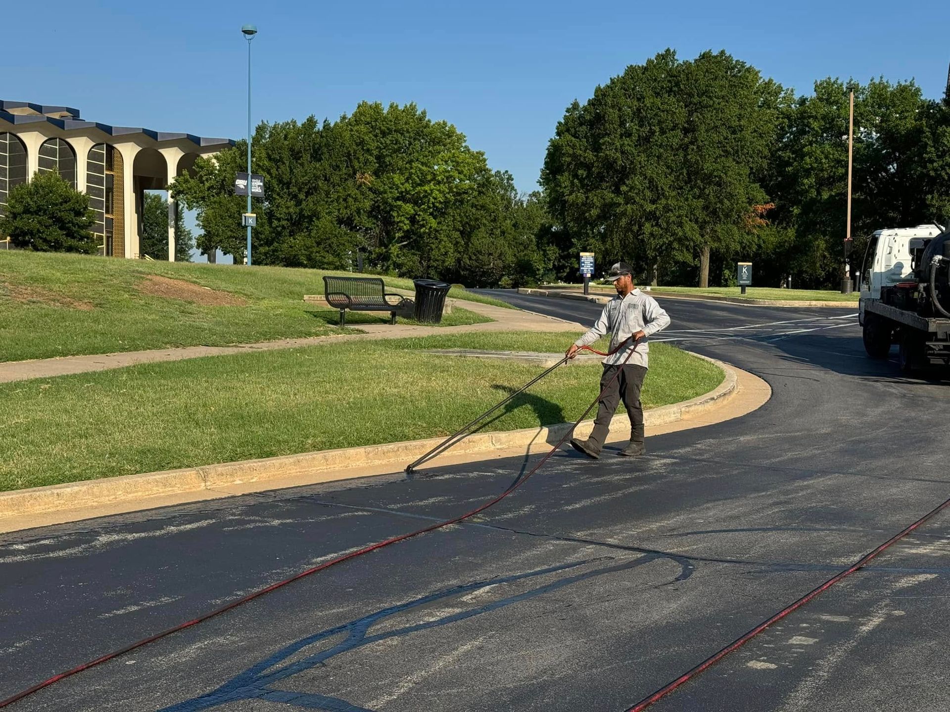 Worker spraying dark sealant on a curved asphalt road beside a grassy park and trees