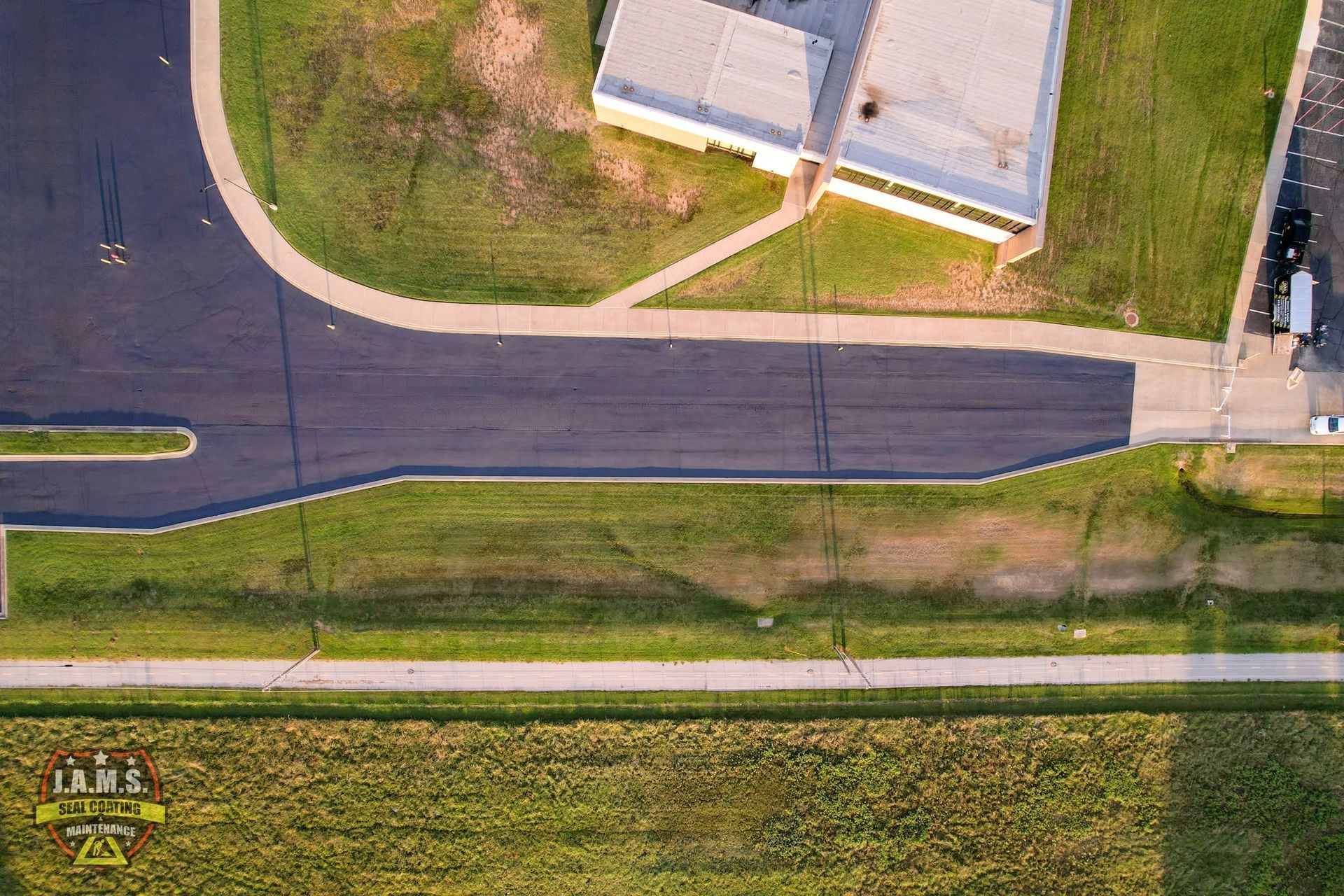 Aerial view of a road beside grassy fields and a building with a sidewalk and driveway.