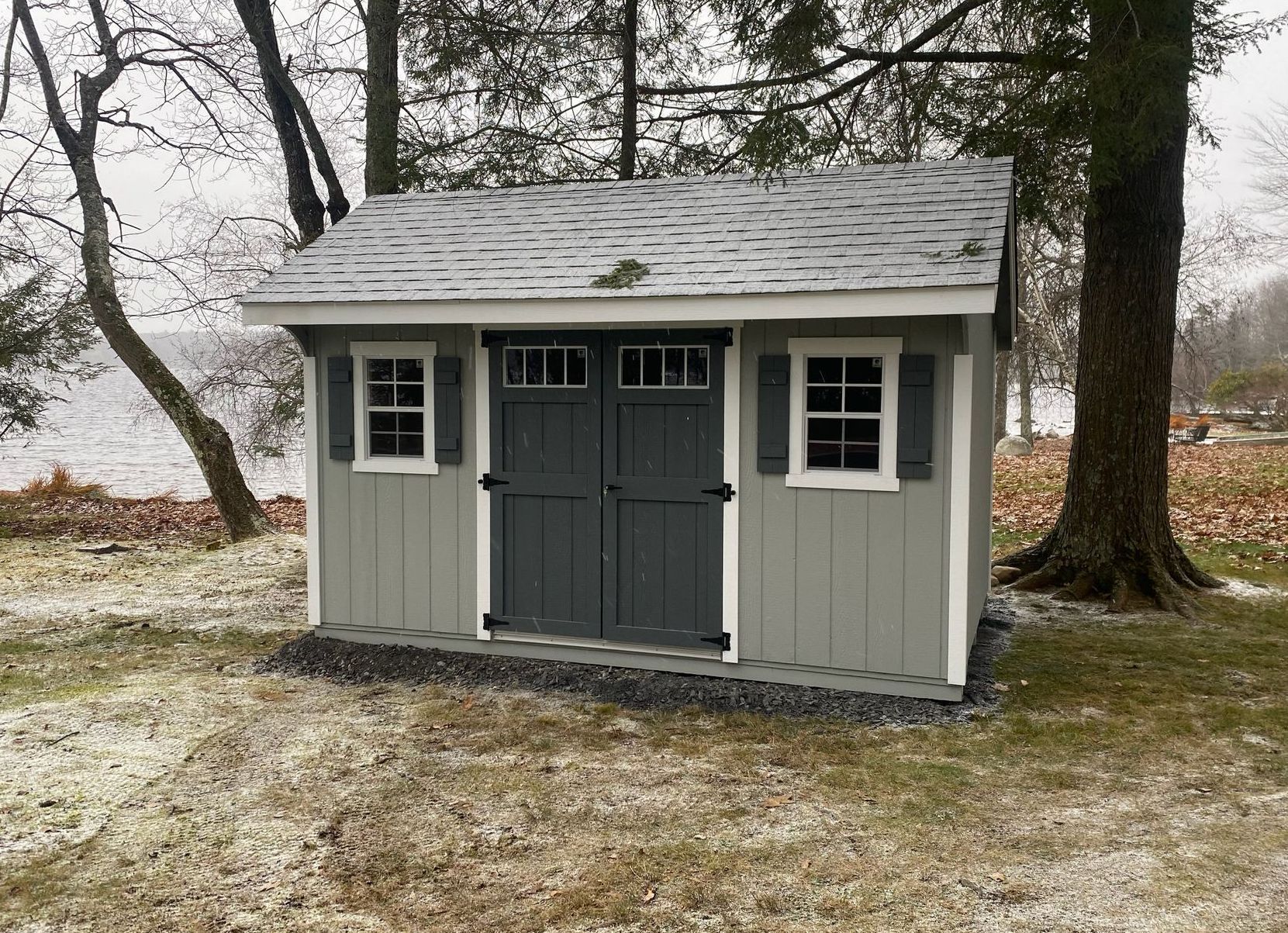 Gray shed with white trim, two windows, and a double door, next to a lake.