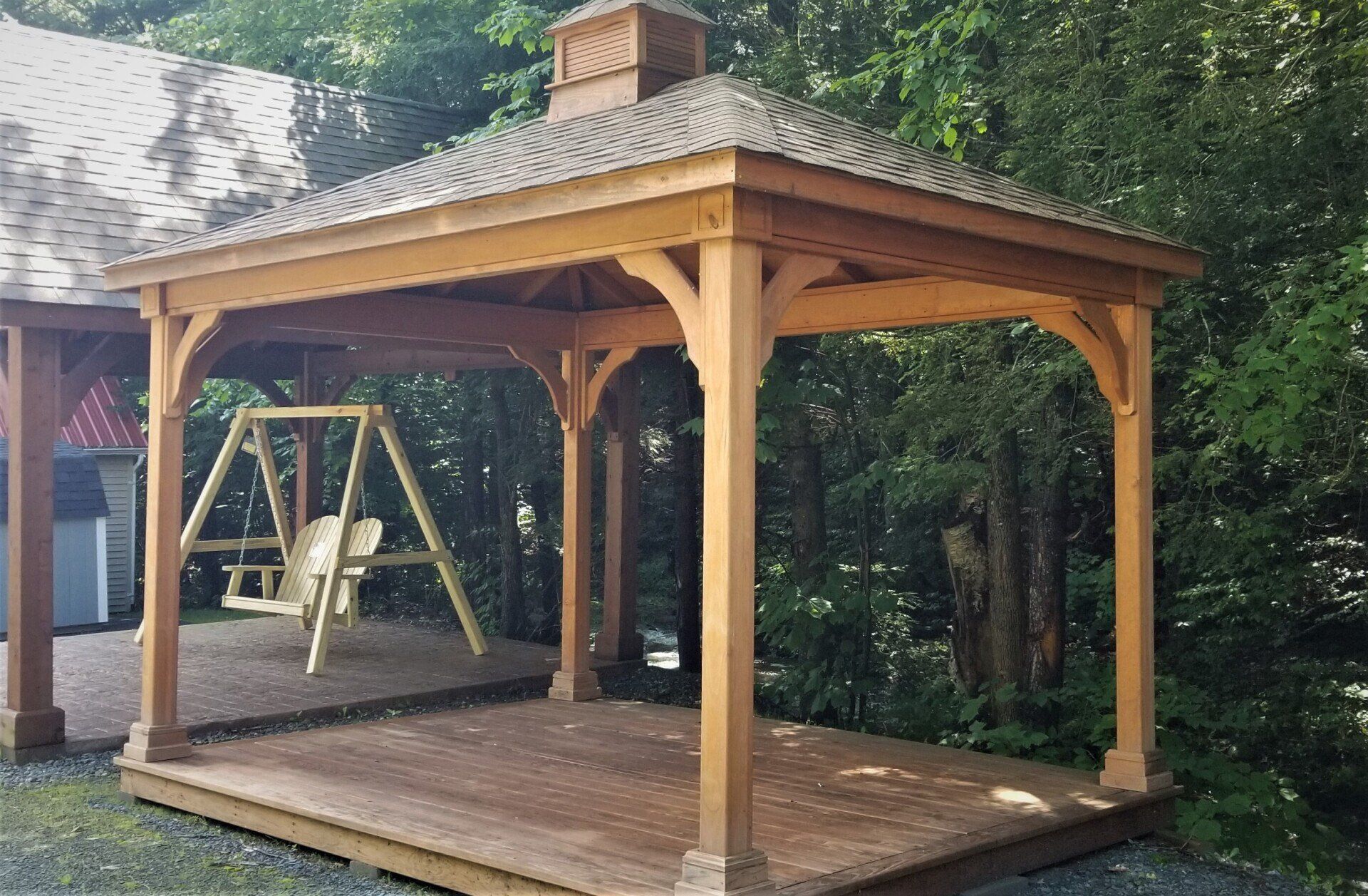 Wooden gazebo with swing on a wooden deck in a wooded area.