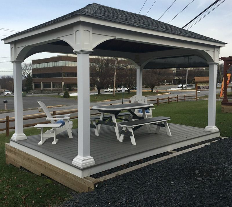 White gazebo with grey deck, outdoor seating: Adirondack chair, picnic table. Building background.
