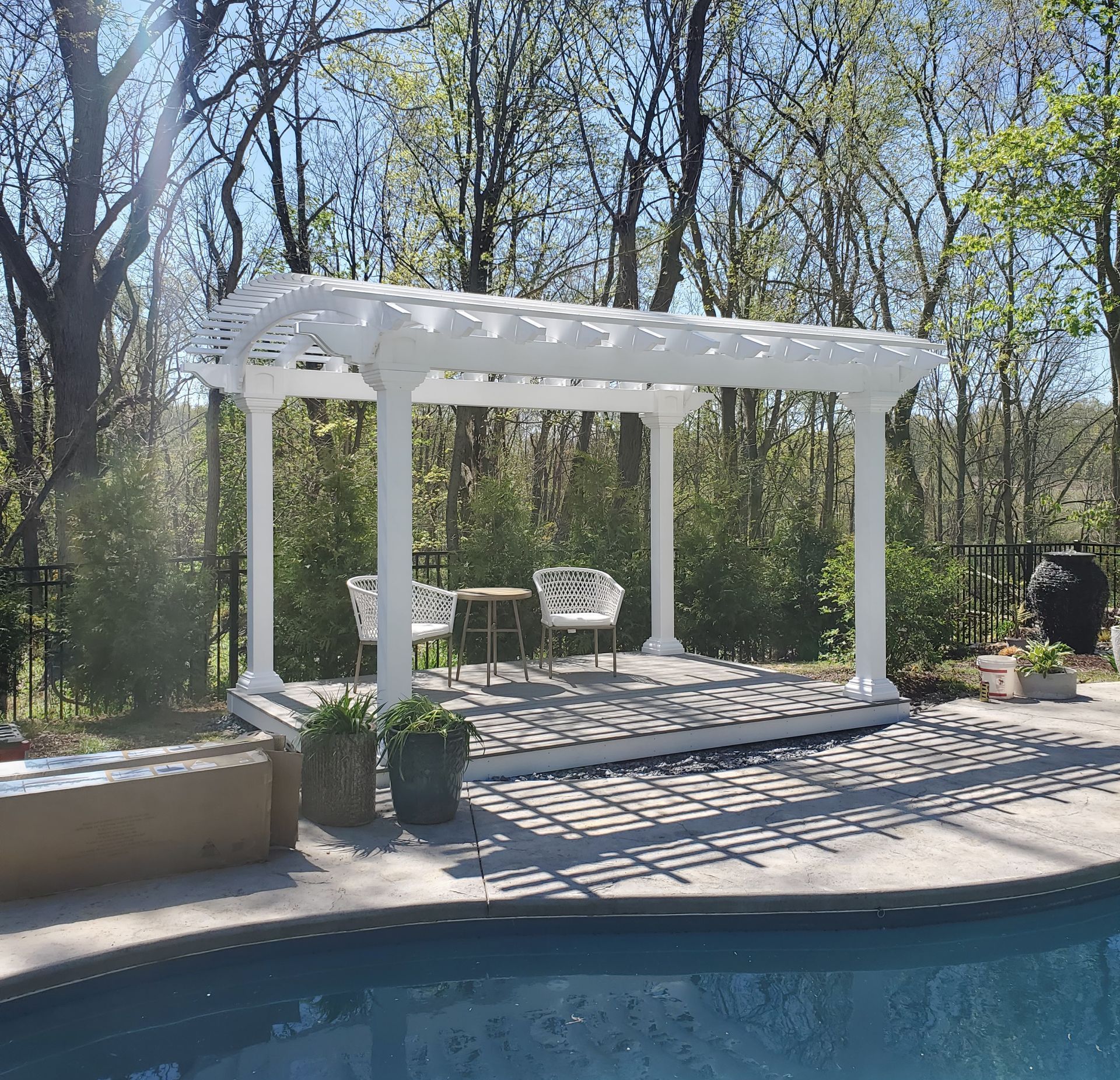 White pergola over a pool, with chairs and a table. Trees in the background. Sunny day.