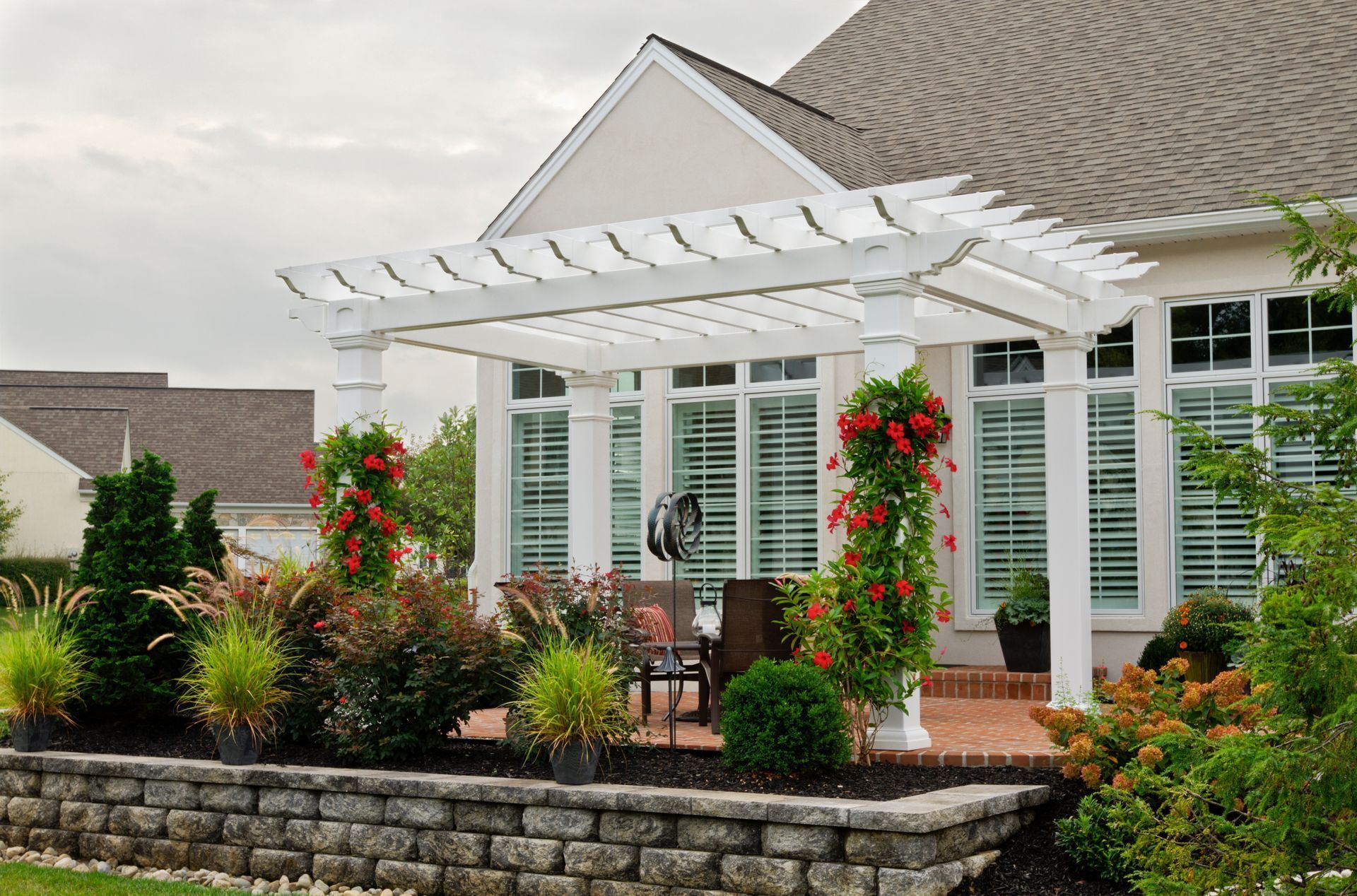 White pergola on a patio with climbing red flowers, overlooking a garden and house.