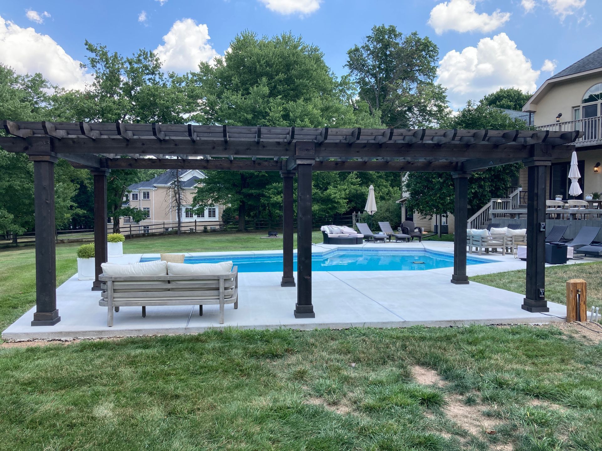 Pergola with seating next to a swimming pool in a backyard on a sunny day.