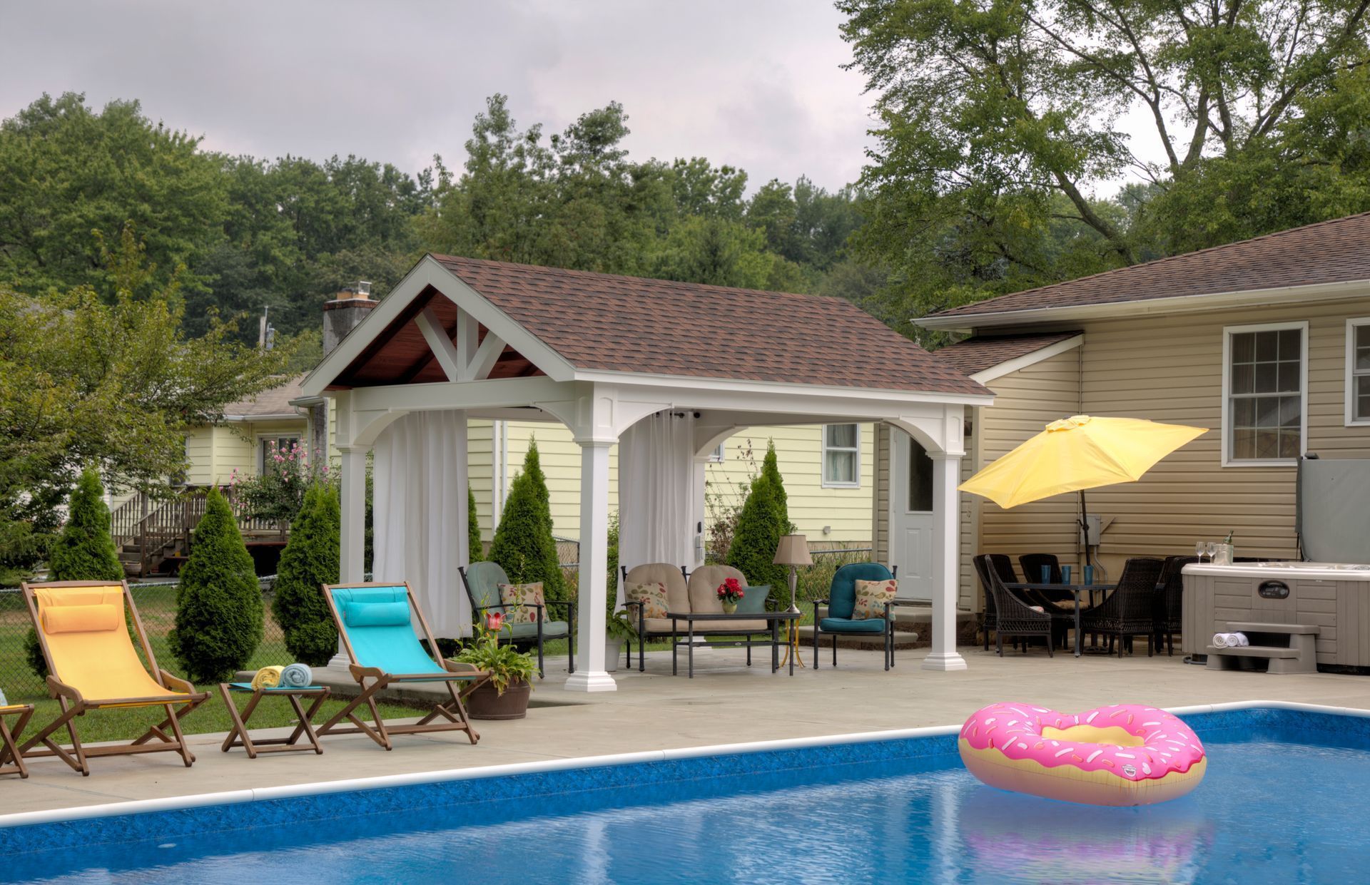 Poolside scene with a gazebo, inflatable donut, chairs, and a yellow umbrella.