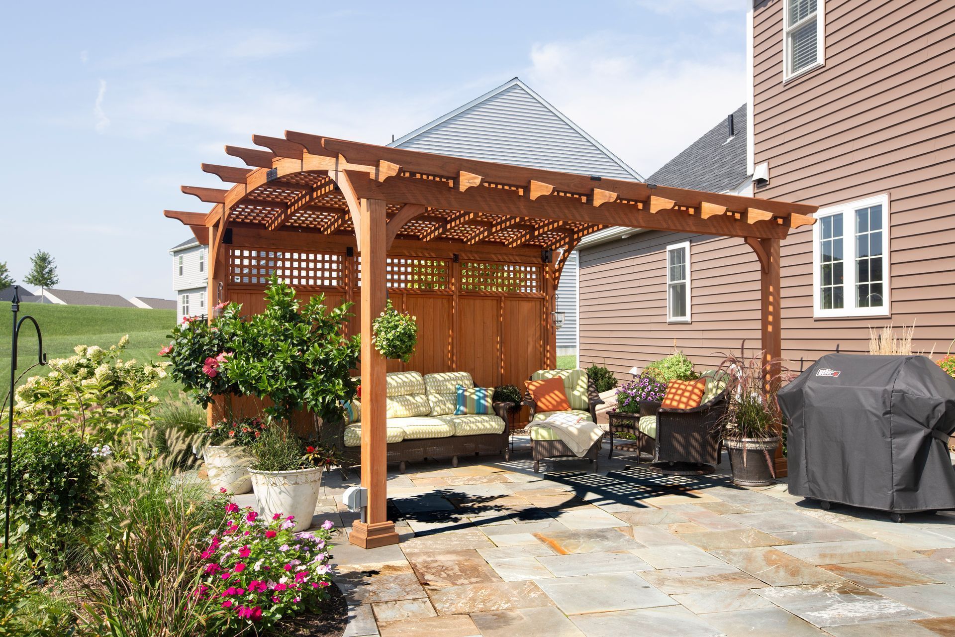 Wooden pergola with seating area in a backyard. Brown wood, stone patio, green plants, and a house in the background.