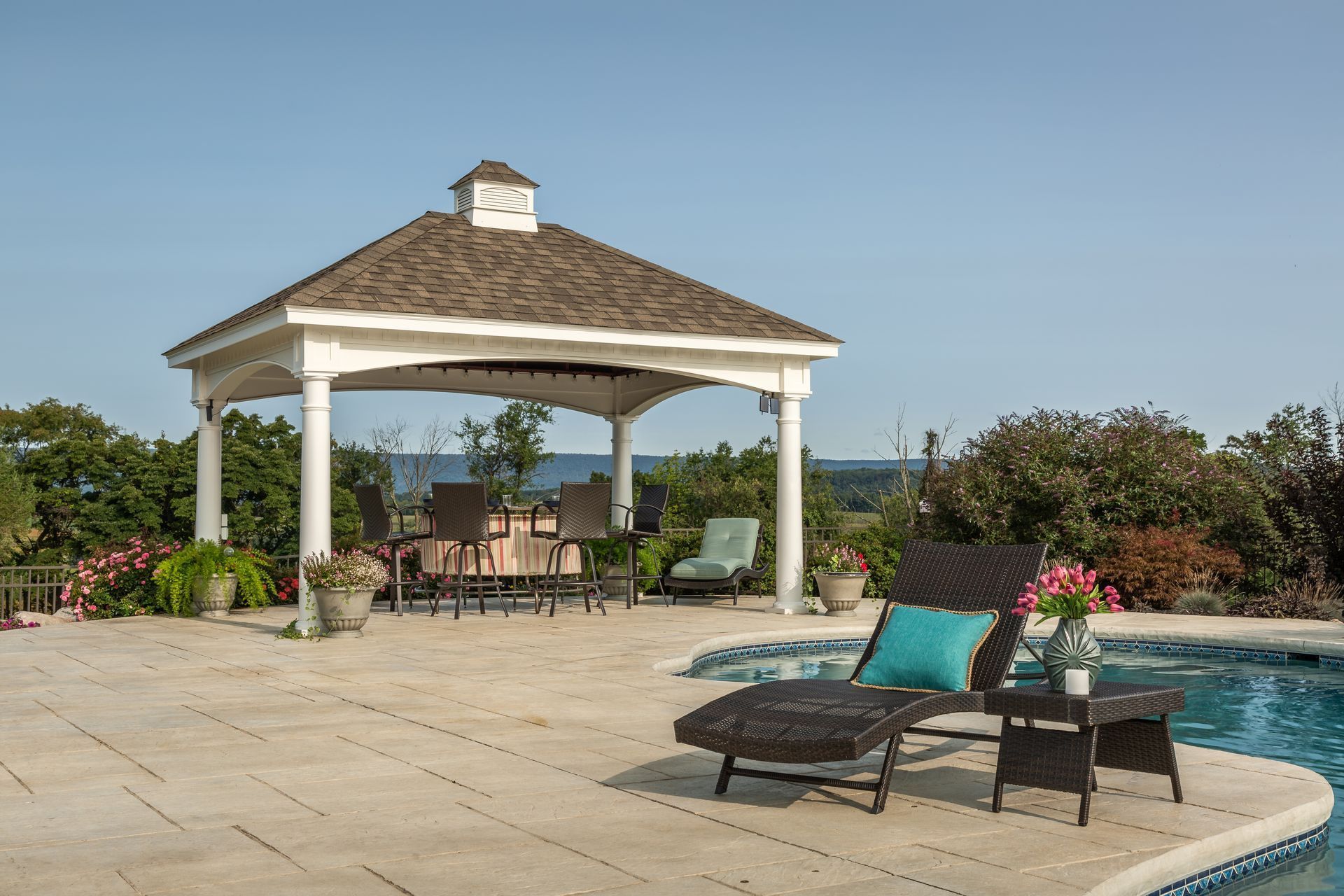 A poolside gazebo with lounge chair, table, and pool under a blue sky.