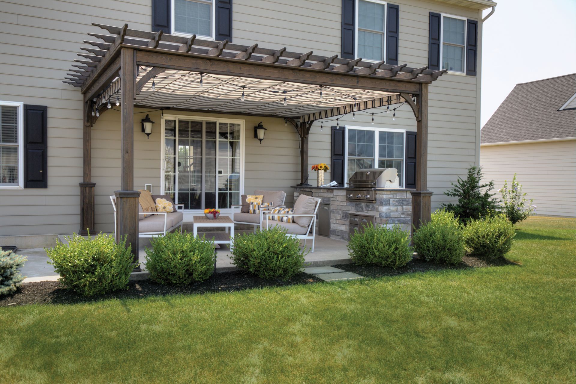 Patio with pergola, seating area, and built-in grill next to a house with green grass.