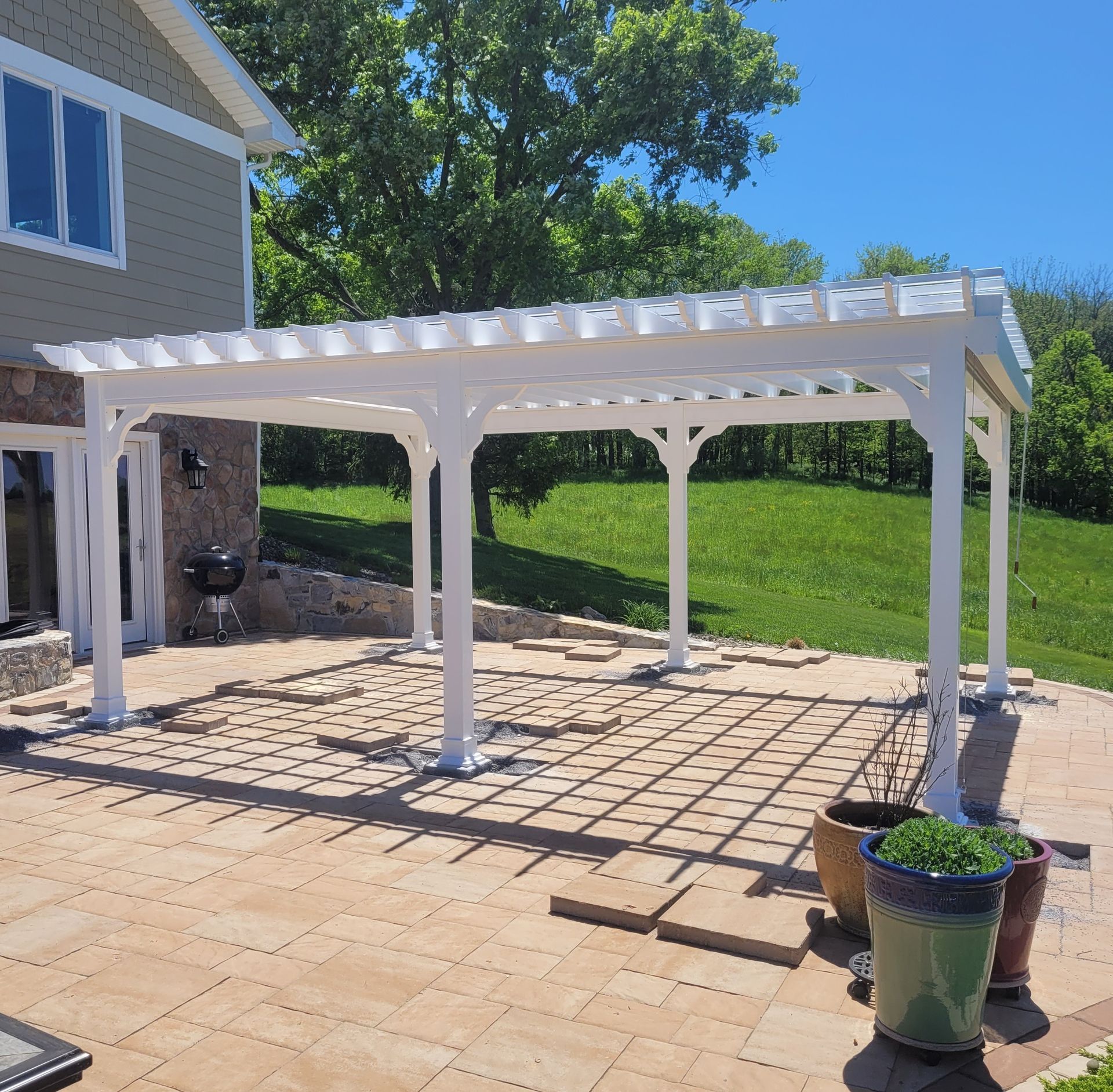 White pergola over a brick patio, near a house, casting shadows on a sunny day.