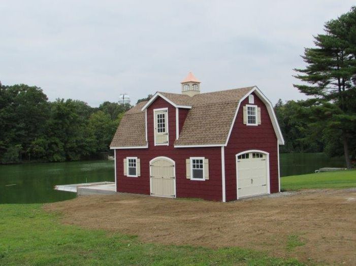 Red boathouse with white trim and tan roof sits near a lake on a grassy lawn.