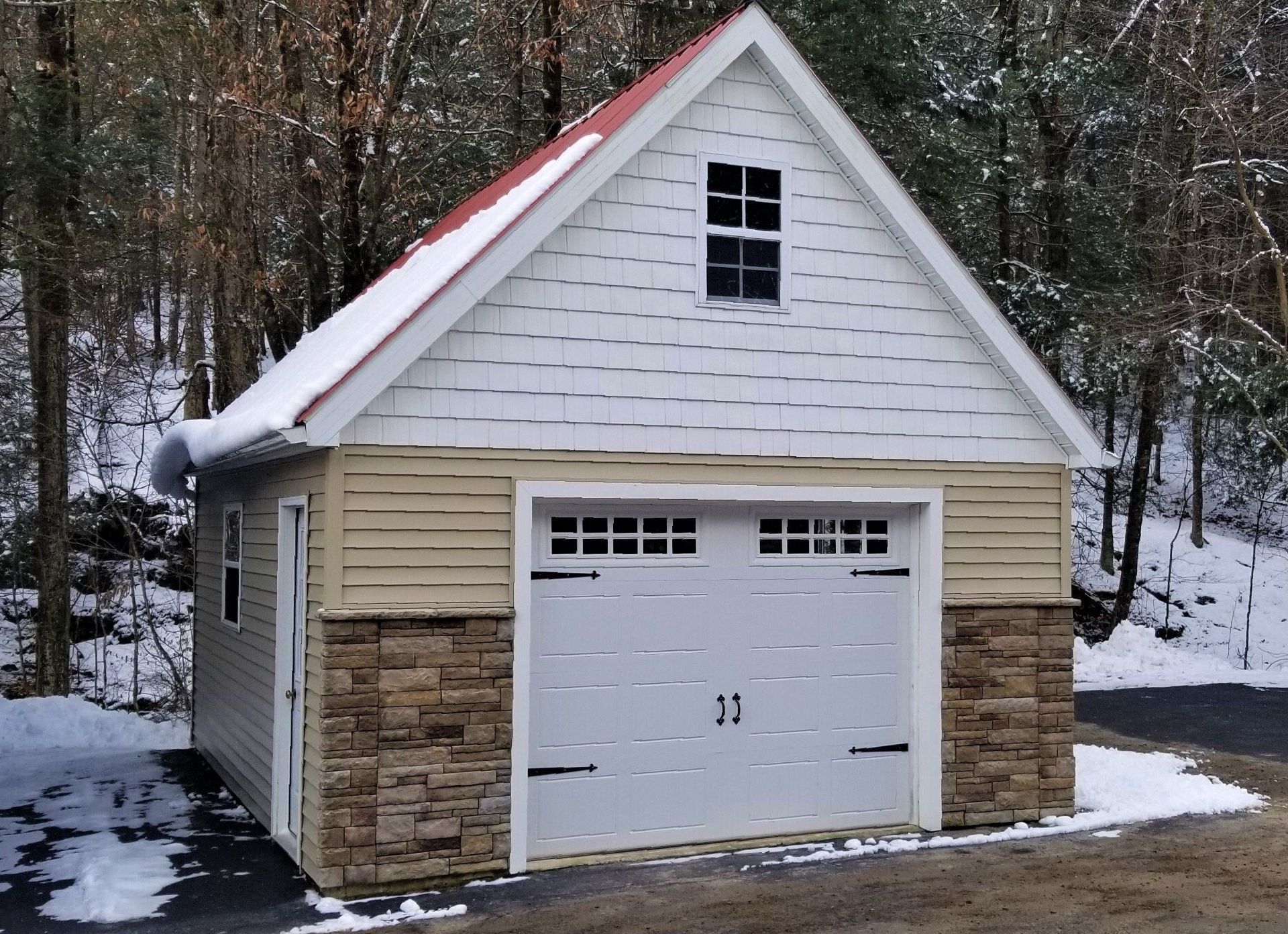 Garage with red roof, stone base, and white garage door in a snowy, wooded area.