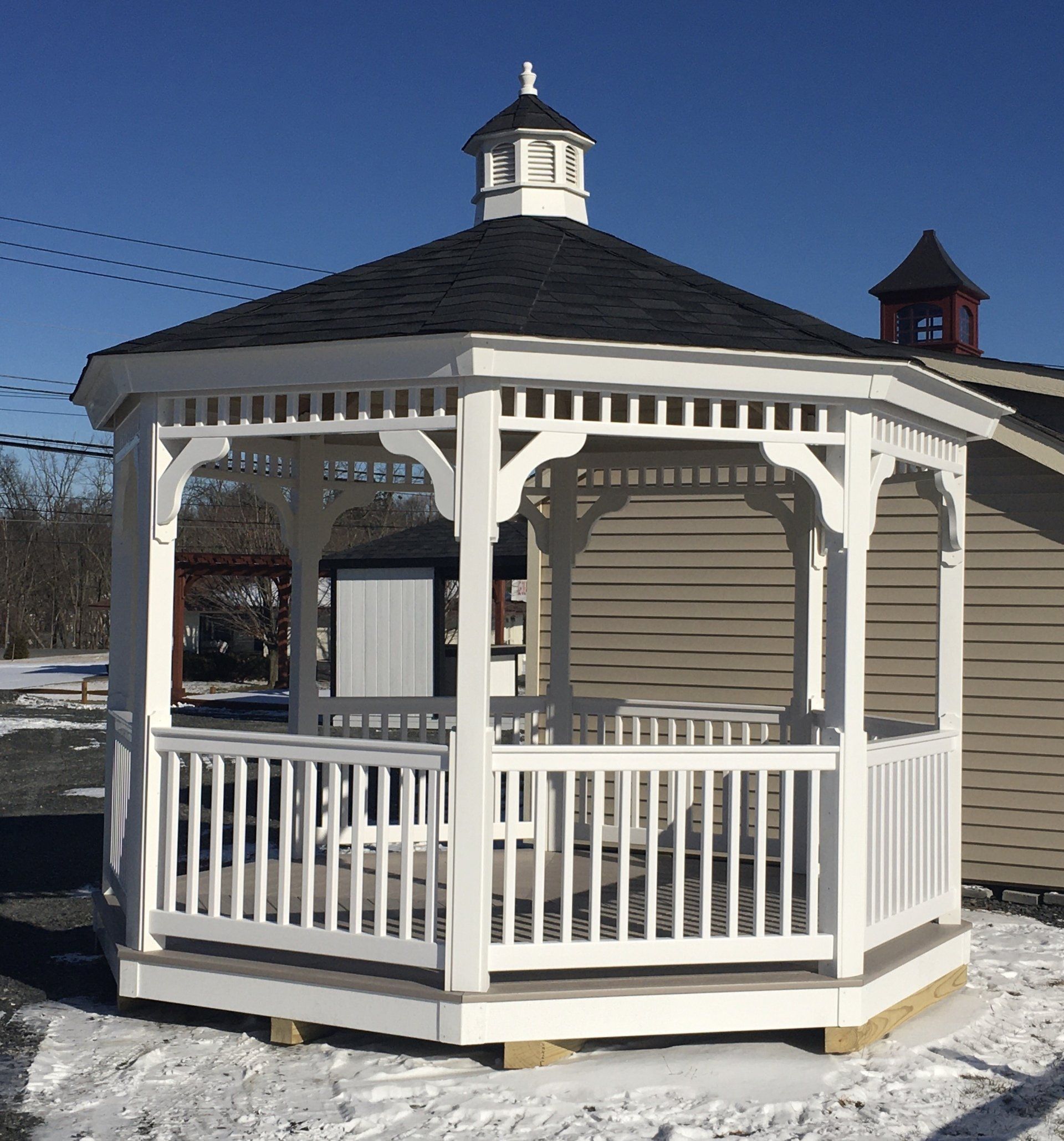 White gazebo with a black roof, set in a snowy area under a clear blue sky.