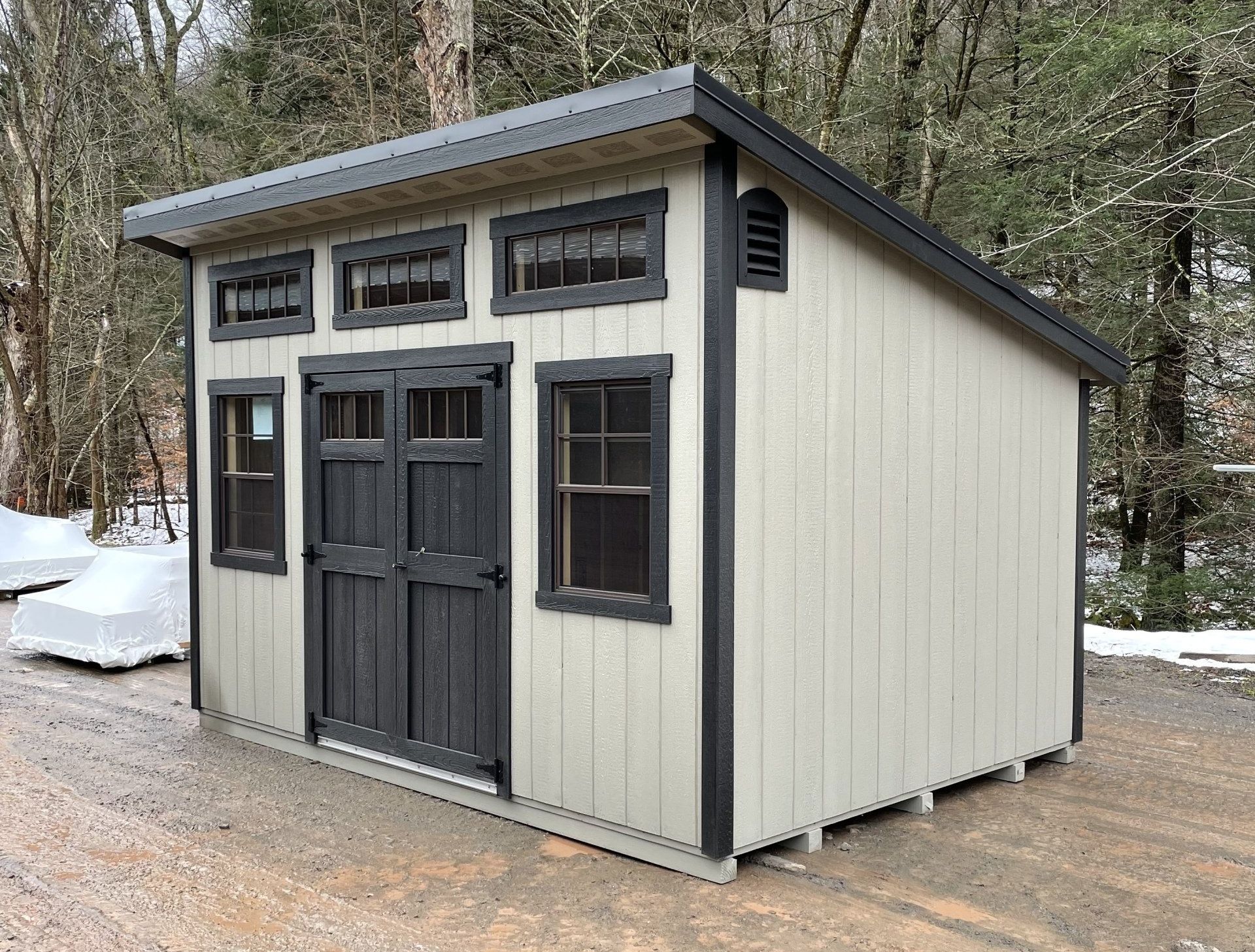 Tan shed with black trim and doors. Windows along the front under a sloped roof, outdoors.