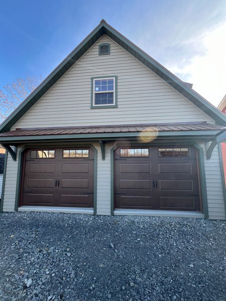Brown garage doors under a tan building with green trim, gravel driveway, blue sky.