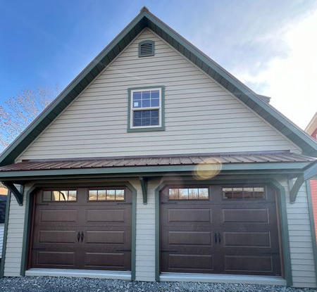 Brown garage doors under a tan building with green trim, gravel driveway, blue sky.