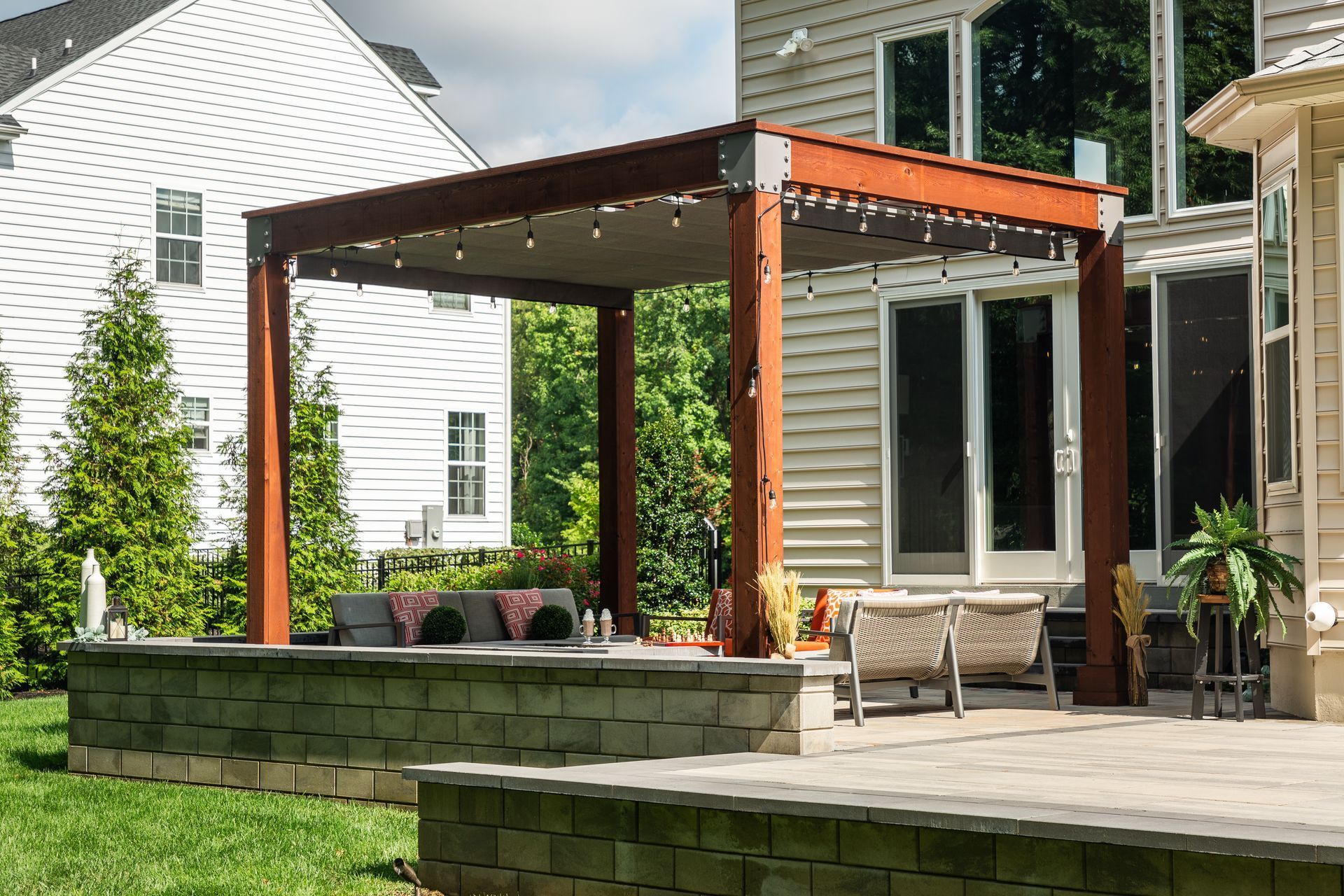 Wooden pergola over stone patio with string lights, next to house.