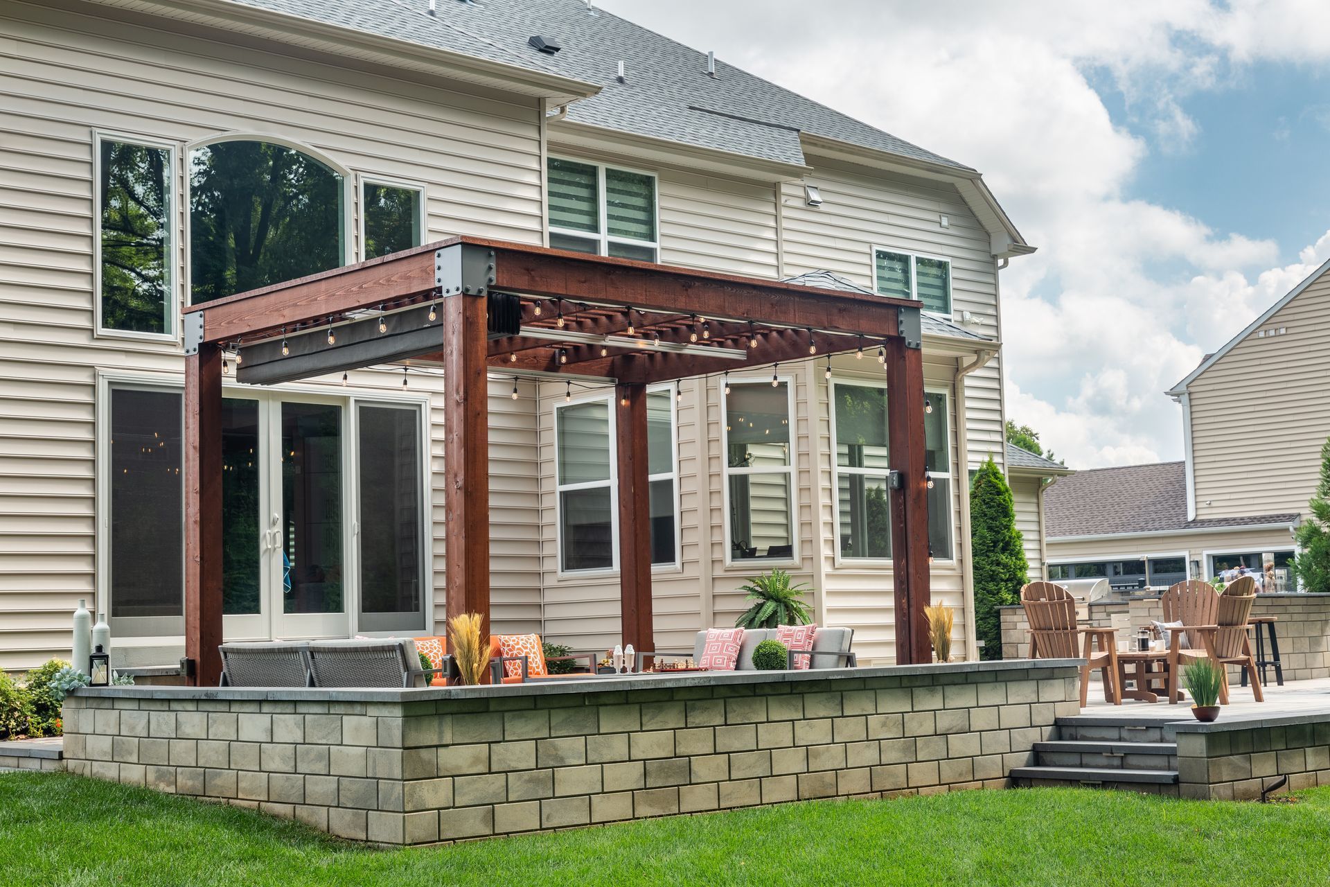 Backyard patio with pergola, beige house, brick retaining wall, and outdoor furniture.