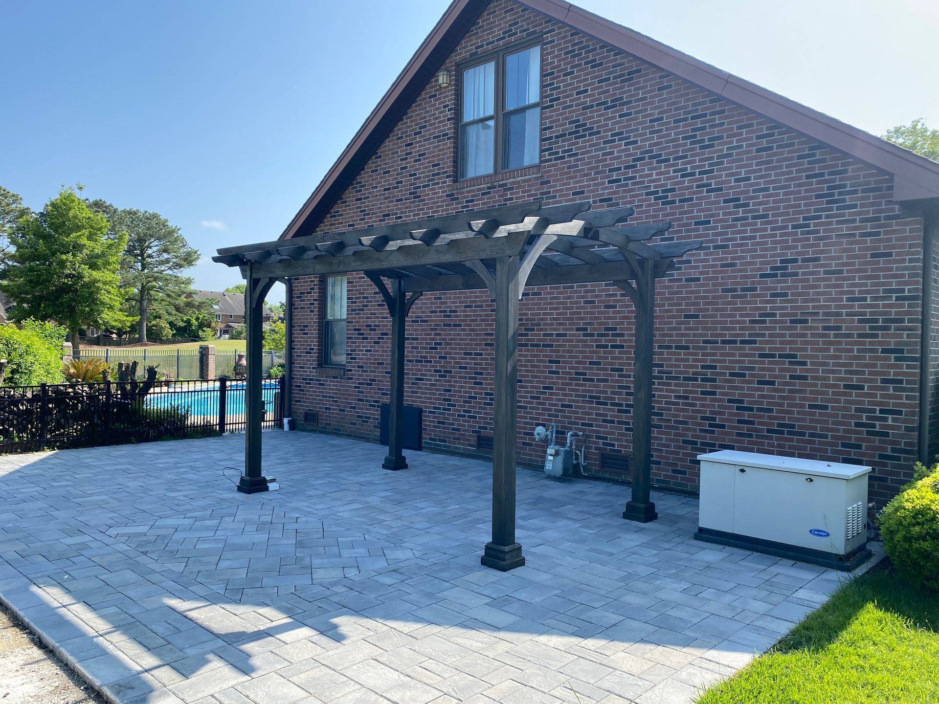 Pergola on a brick patio, next to a house. Pool in background.